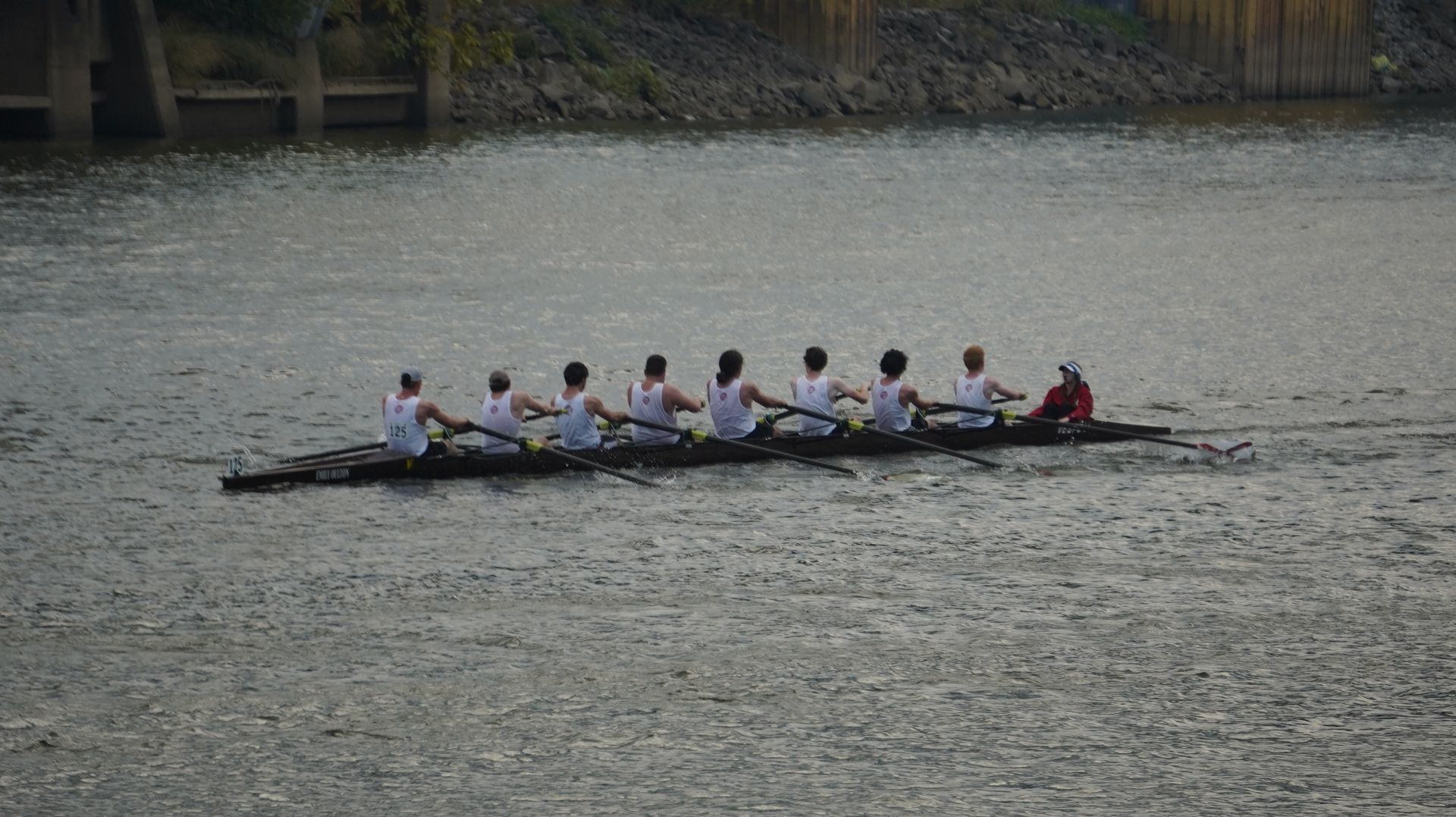Rowing team on a river, pulling oars in unison. White tank tops and dark boat against a gray-green water backdrop.