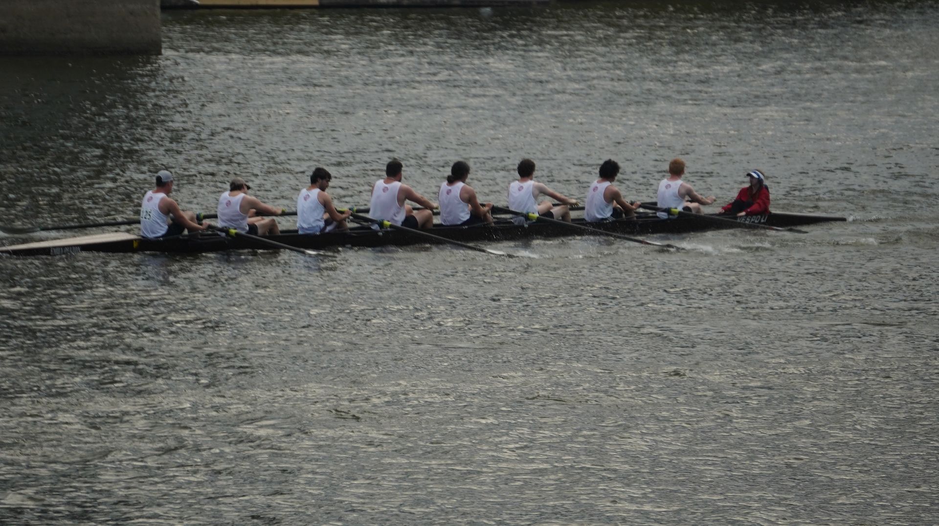 Rowing team in a long boat on a river, eight rowers facing backward, white shirts, dark boat.