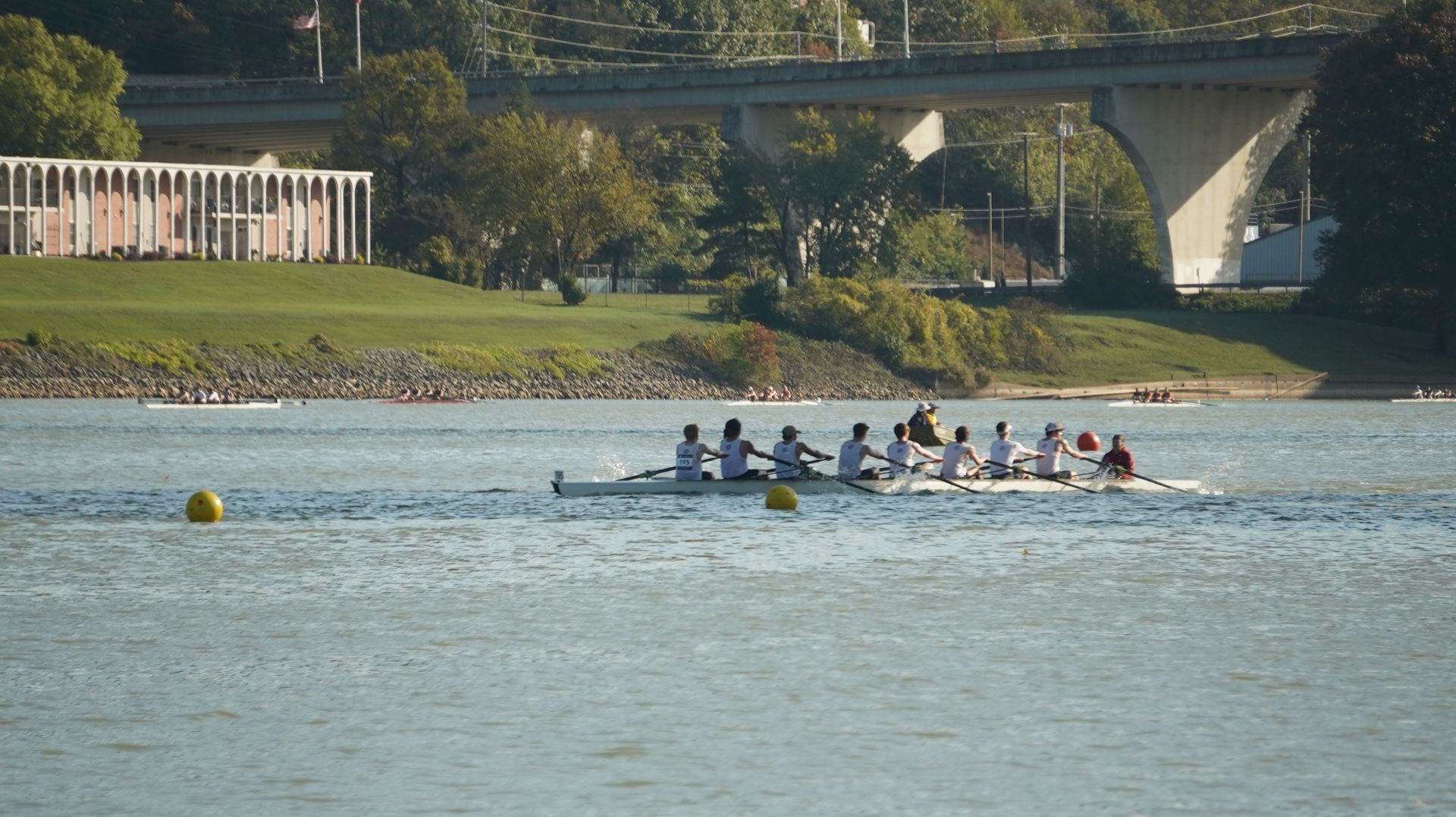 A rowing team in a long boat on a river with a bridge in the background. They are wearing white and blue.