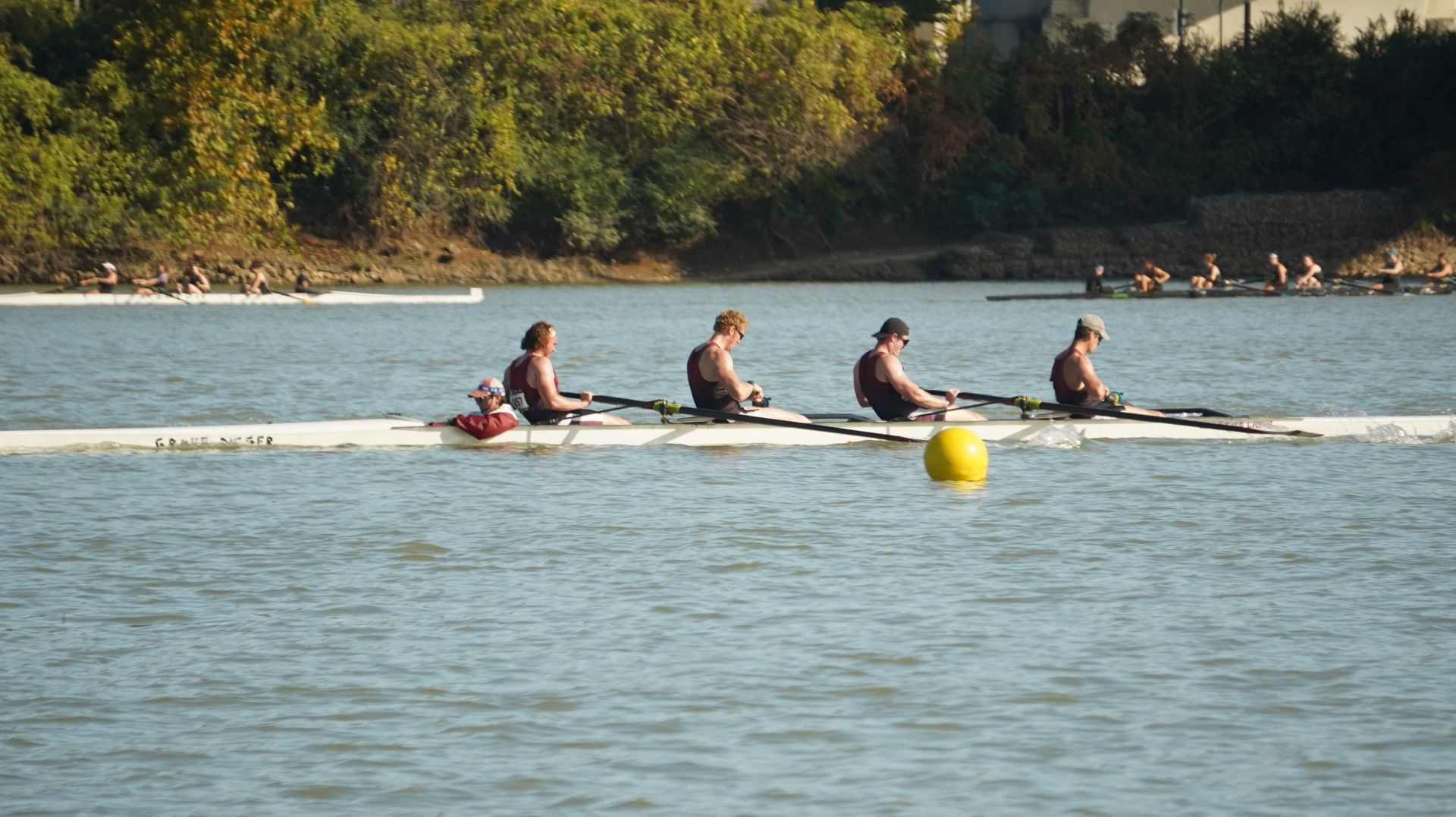 Four rowers on a long white boat on a river, with two other boats in the distance and a yellow buoy.