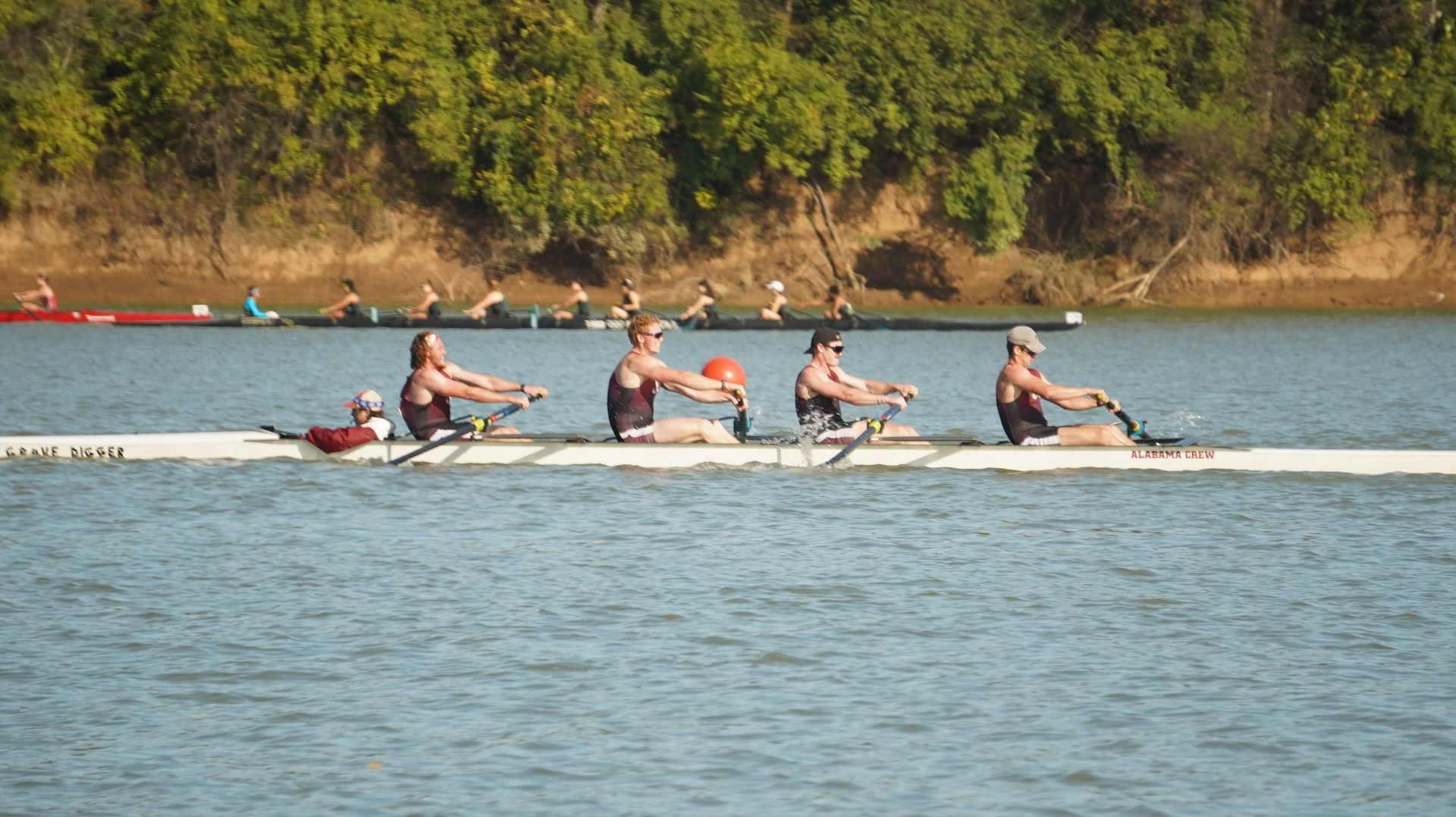 Four rowers on a long, narrow boat on a river, with a second boat in the background. Sunlight glints on the water.