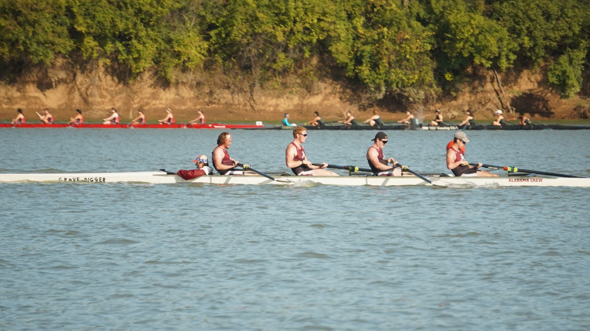 Rowing race on a river; multiple teams competing, including one in the foreground. Trees line the riverbank on a sunny day.
