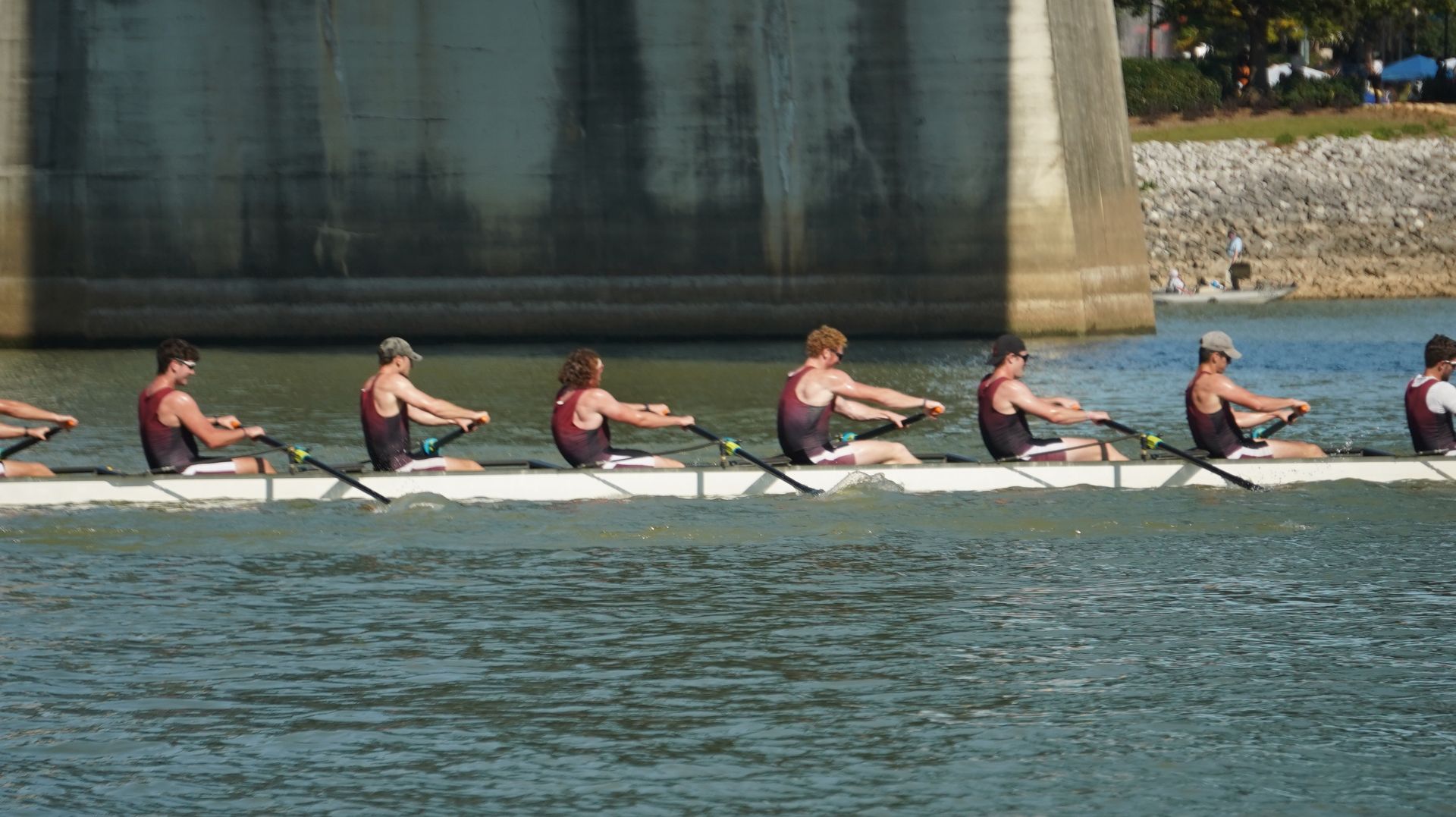 Rowing team in a long white boat on a river, under a bridge; athletes in maroon and black uniforms rowing in unison.
