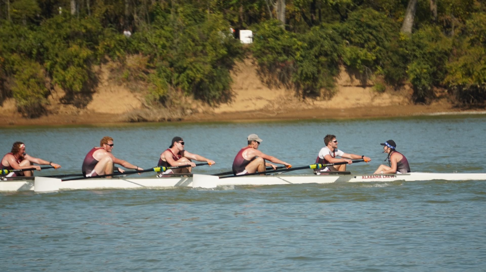 Rowing crew in a white boat on a river, rowing towards the viewer. Trees and a dirt bank are in the background.