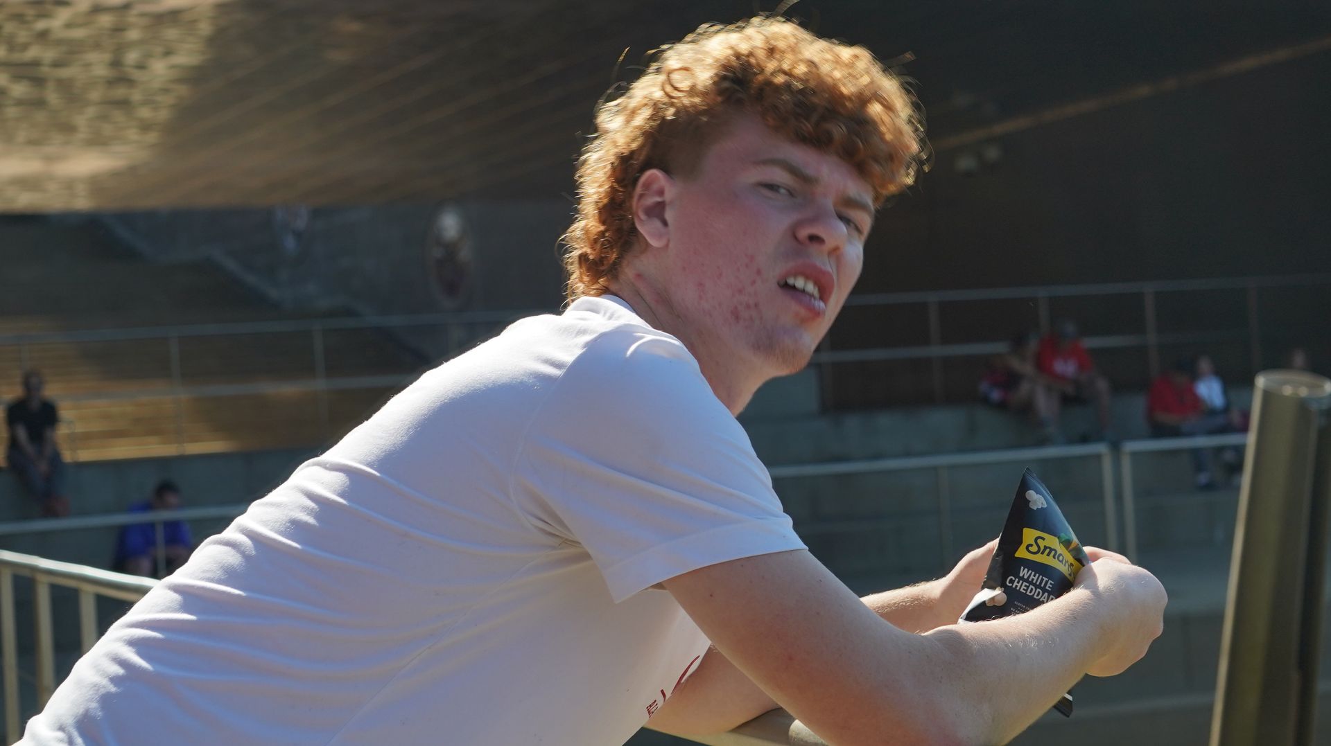 A young man with red, curly hair leans on a railing, looking upward with a slight frown, holding a snack package outdoors.
