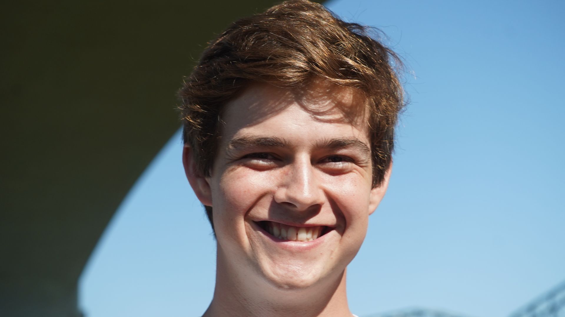 Young man with light brown hair smiles broadly at the camera against a blue sky.