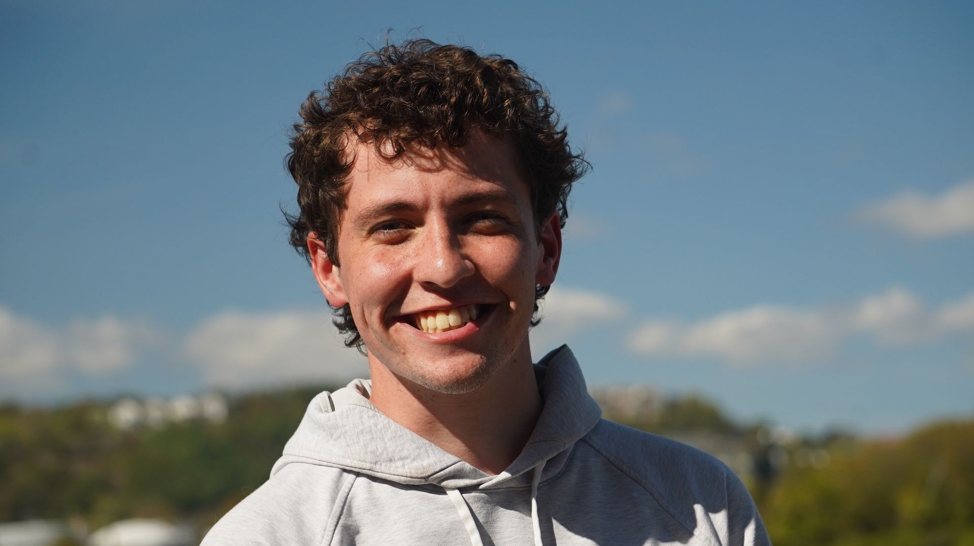 A smiling young man with curly brown hair, wearing a grey hoodie, stands outdoors on a sunny day.