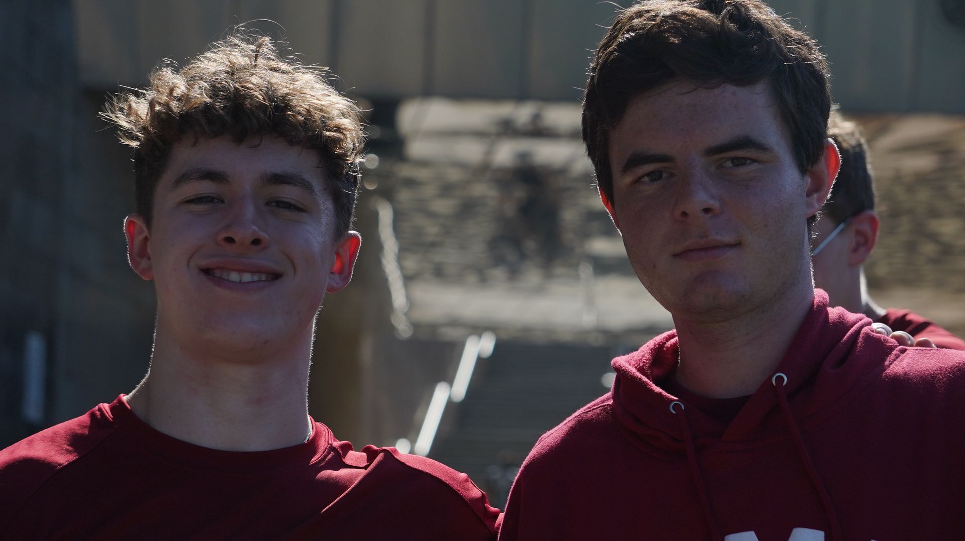 Two young men smiling, wearing maroon shirts, posing outdoors in front of a building.
