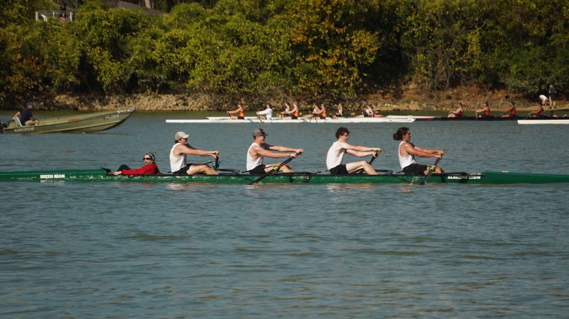 A rowing team in a long green boat on a river, trees in the background. Five rowers wearing white and black actively rowing.