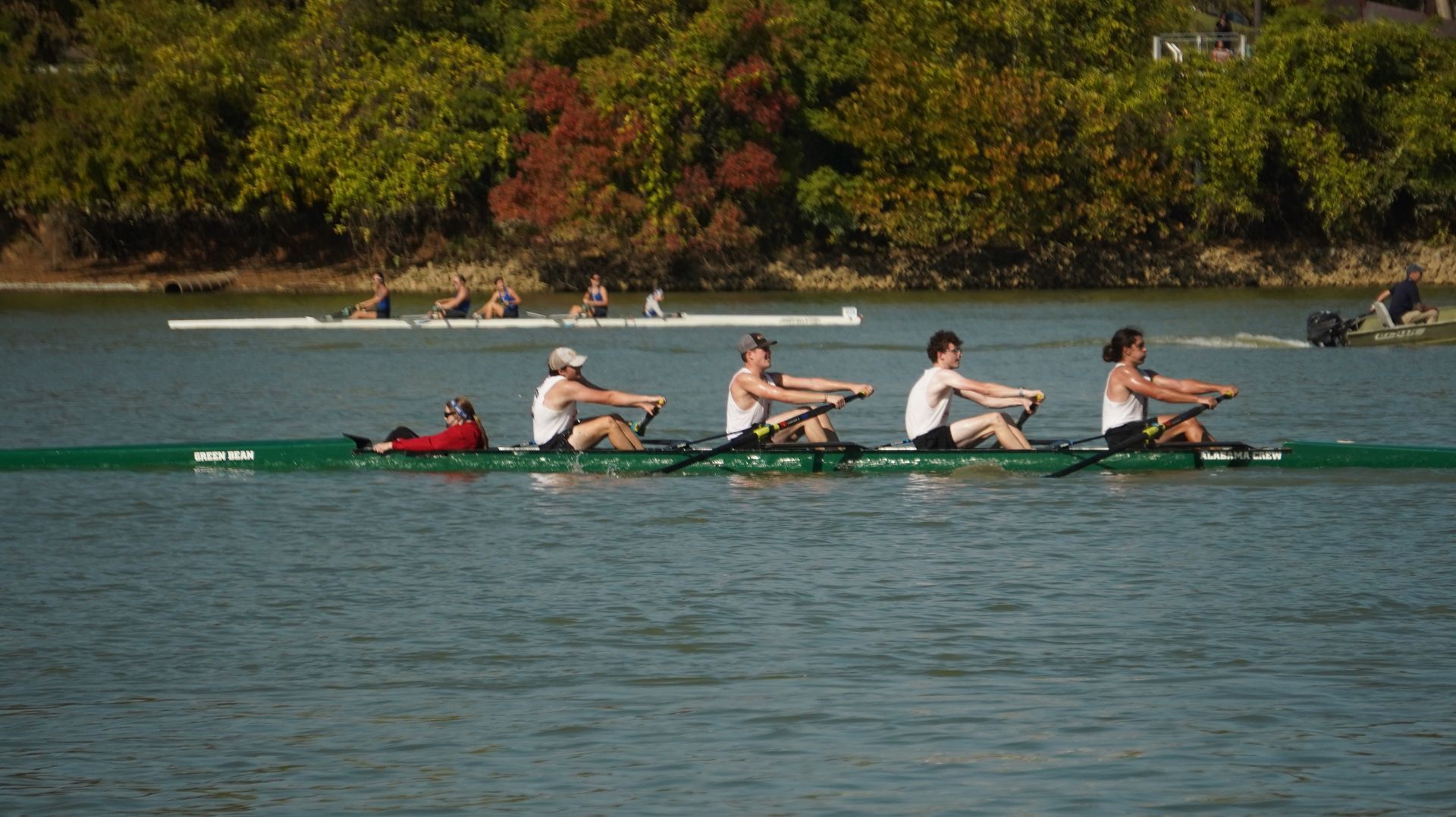 A rowing team in a green boat on a lake, with a coach lying down, trees in the background.