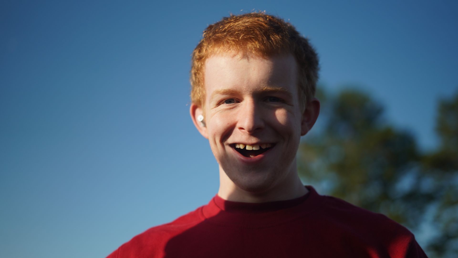 A young man with red hair smiles at the camera, set against a clear blue sky. He wears a red shirt and has an earbud in.