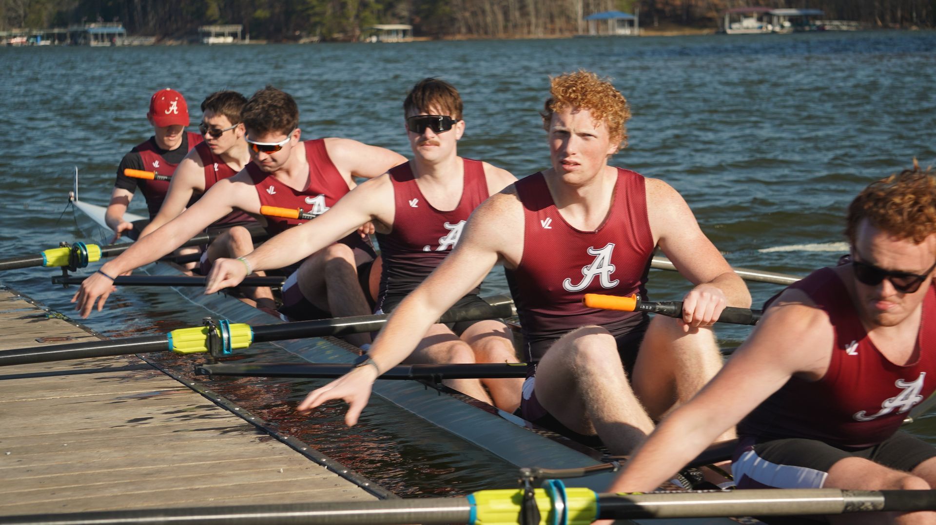 Rowing team in maroon and black uniforms on a lake, rowing in unison. The men are focused and determined.