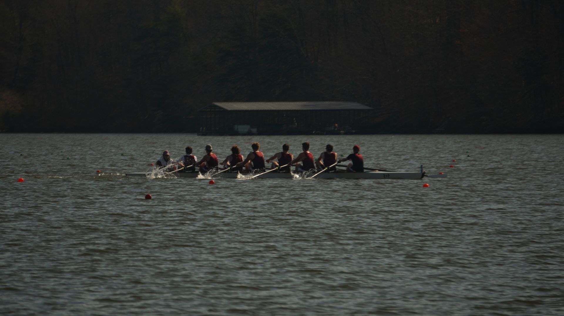 A rowing team on a lake, with eight people in a long boat, dark water, and a treeline in the background.
