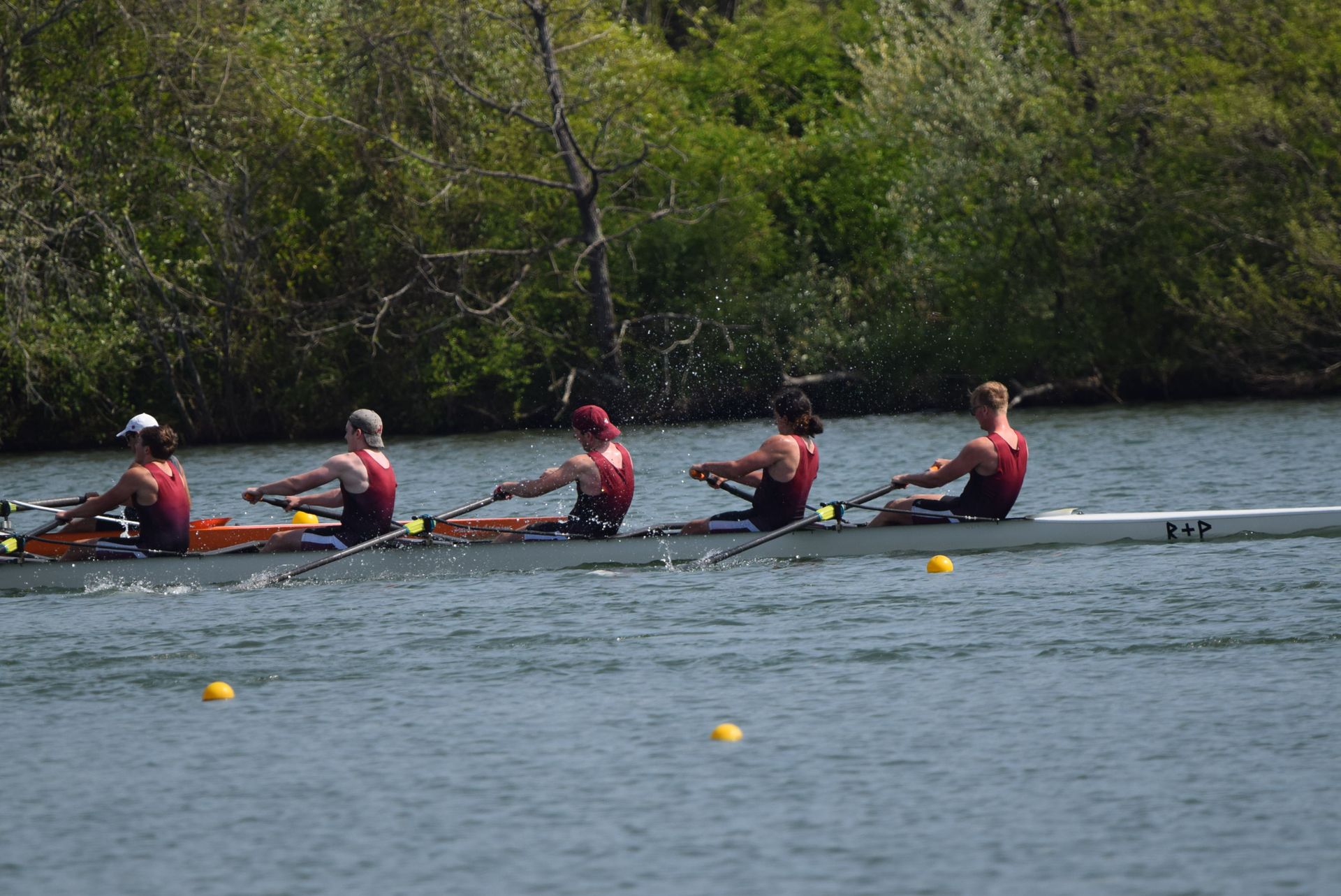 A rowing team in maroon uniforms competes on a river, trees line the bank, and buoys mark the course.