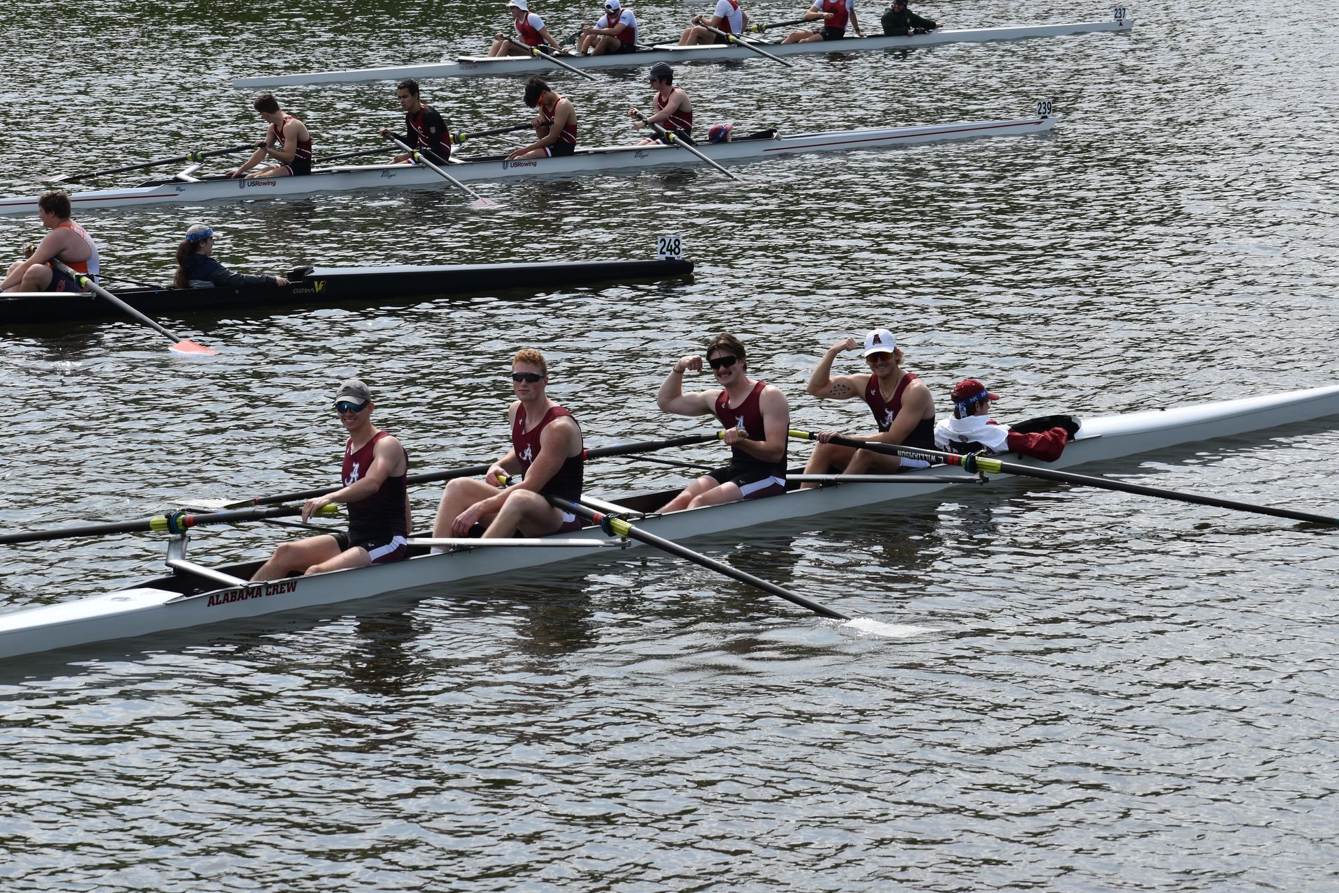 Rowing crew in a long boat on water, arms raised in victory. Multiple boats visible, competing on a river.