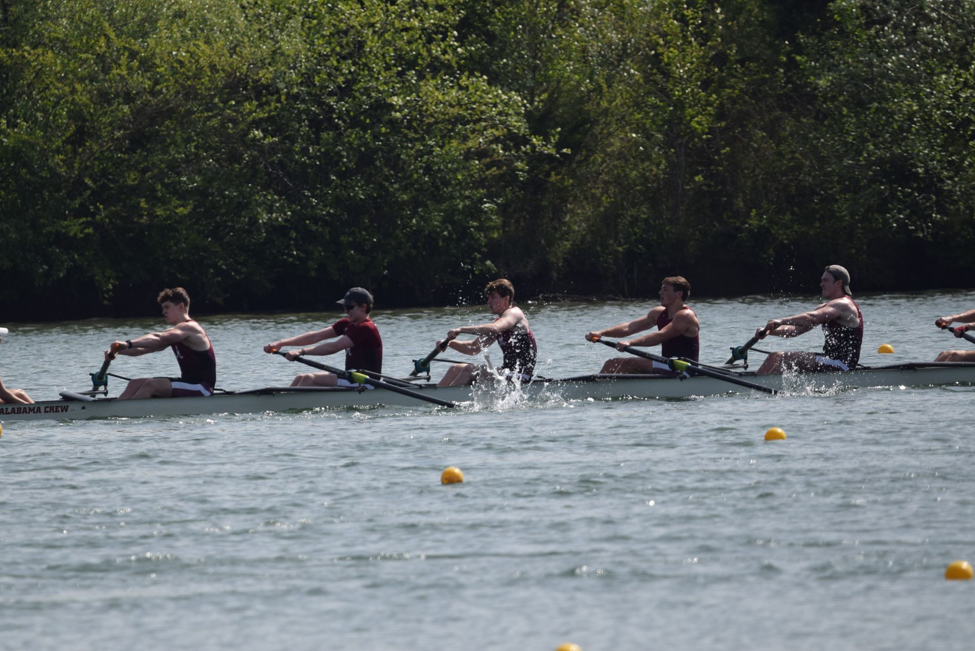Rowing crew racing on a waterway. Five men in a long, narrow boat are actively rowing, splashing water.