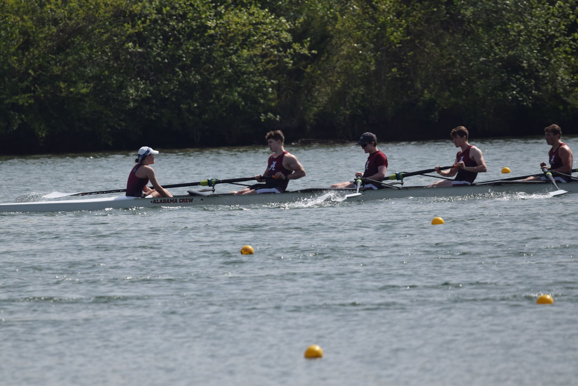 Rowing team in a long boat on a river. Five people wearing burgundy uniforms are rowing; a coxswain in a white hat steers.