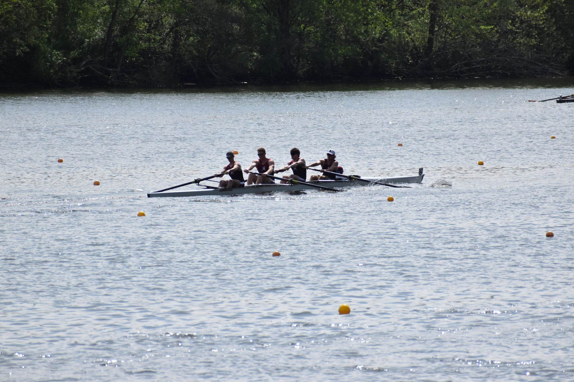 Four rowers in a boat on a lake, rowing in sync. They are wearing dark clothing. Yellow buoys float in the water.