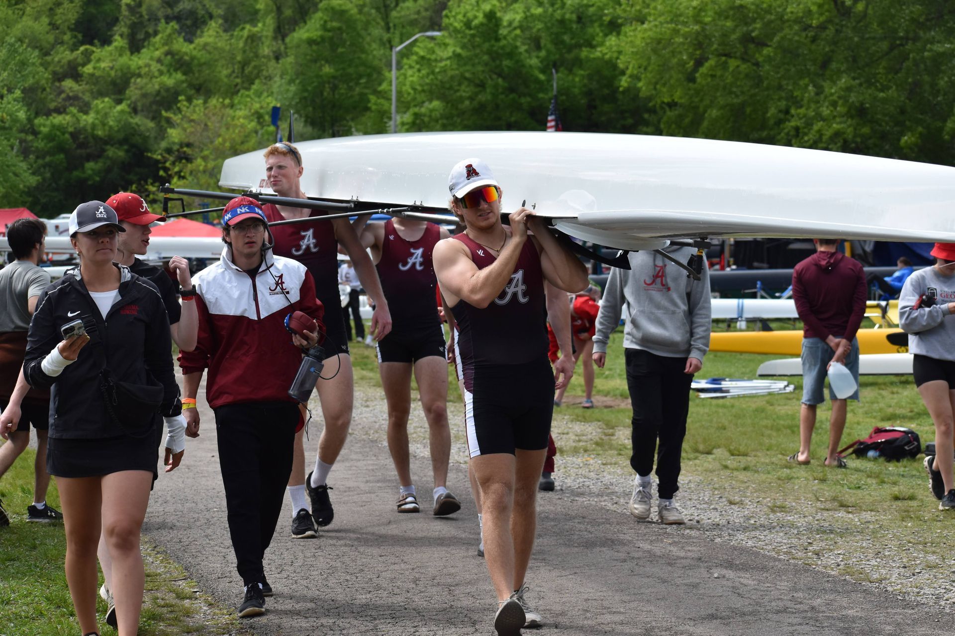 A group of rowers in maroon and white carrying a boat over their heads on a dirt path.