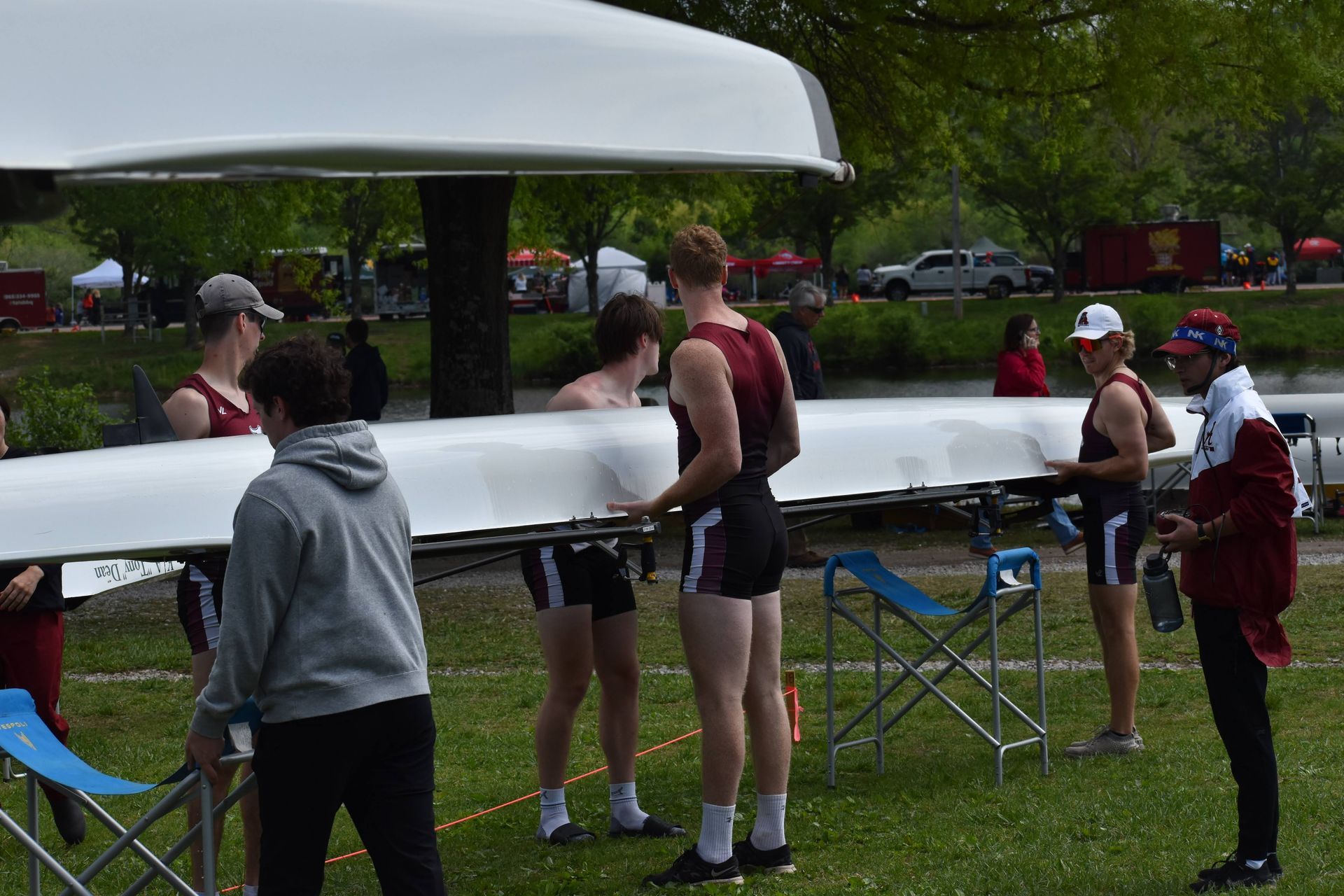 Rowing team members preparing a long white boat on the grass near a body of water; others in red uniforms watch.