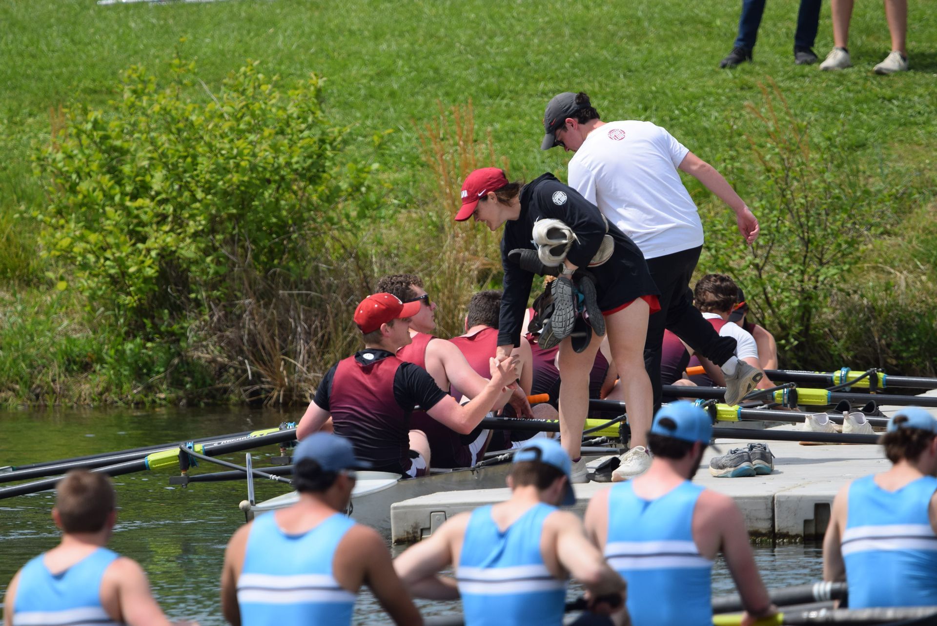 Rowing team members assist each other near the water's edge. A coach in a white shirt and cap is also present.