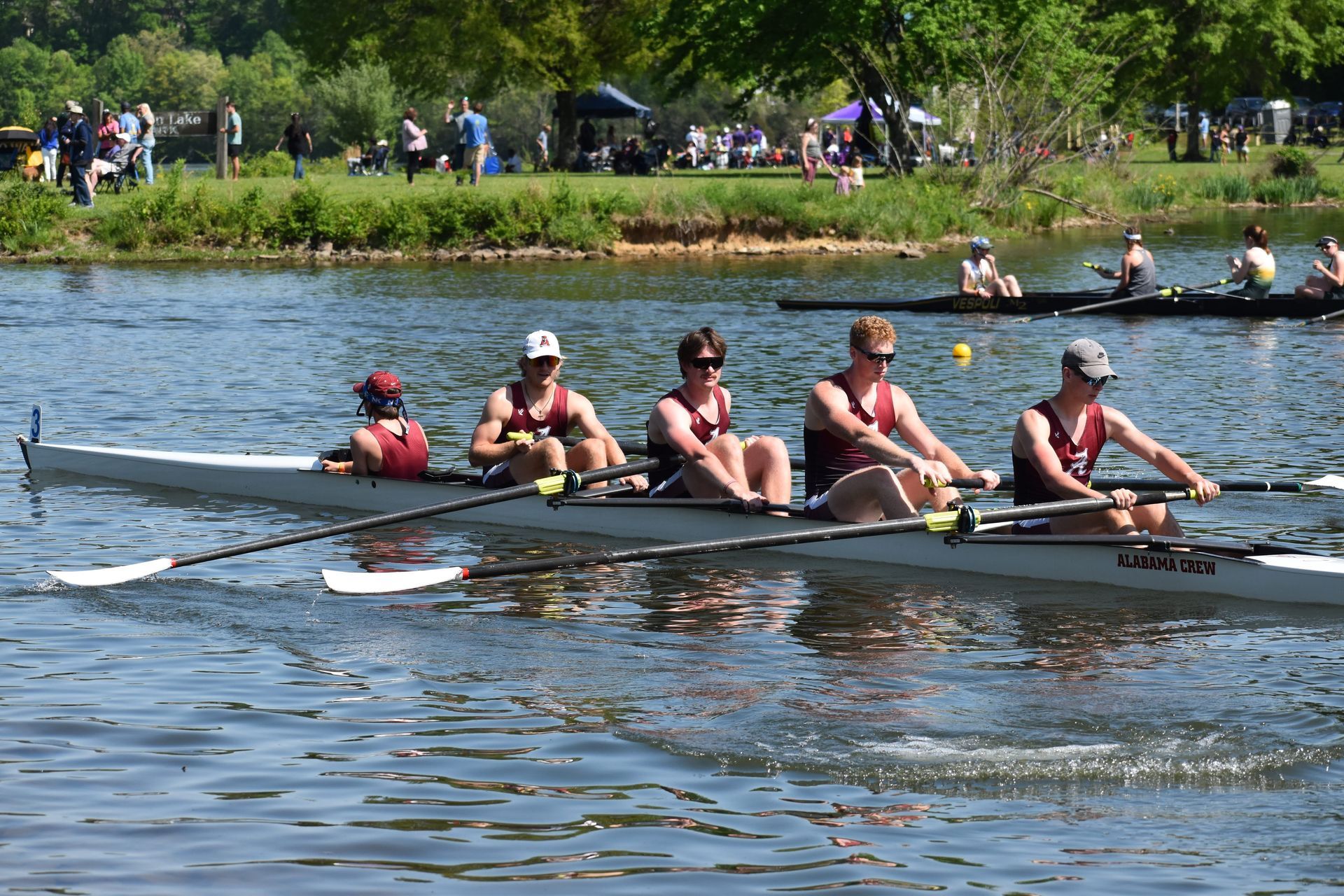 A rowing team in a long boat on a lake, with four rowers facing backward, pulling oars. Spectators watch from the shore.