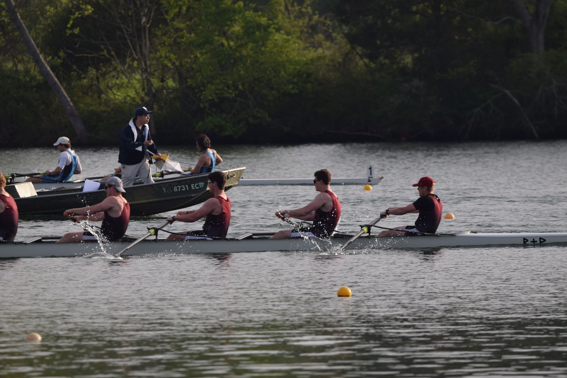 Rowing crew on the water, two boats racing. The rowers wear maroon and white, with a coach following in a motorboat.