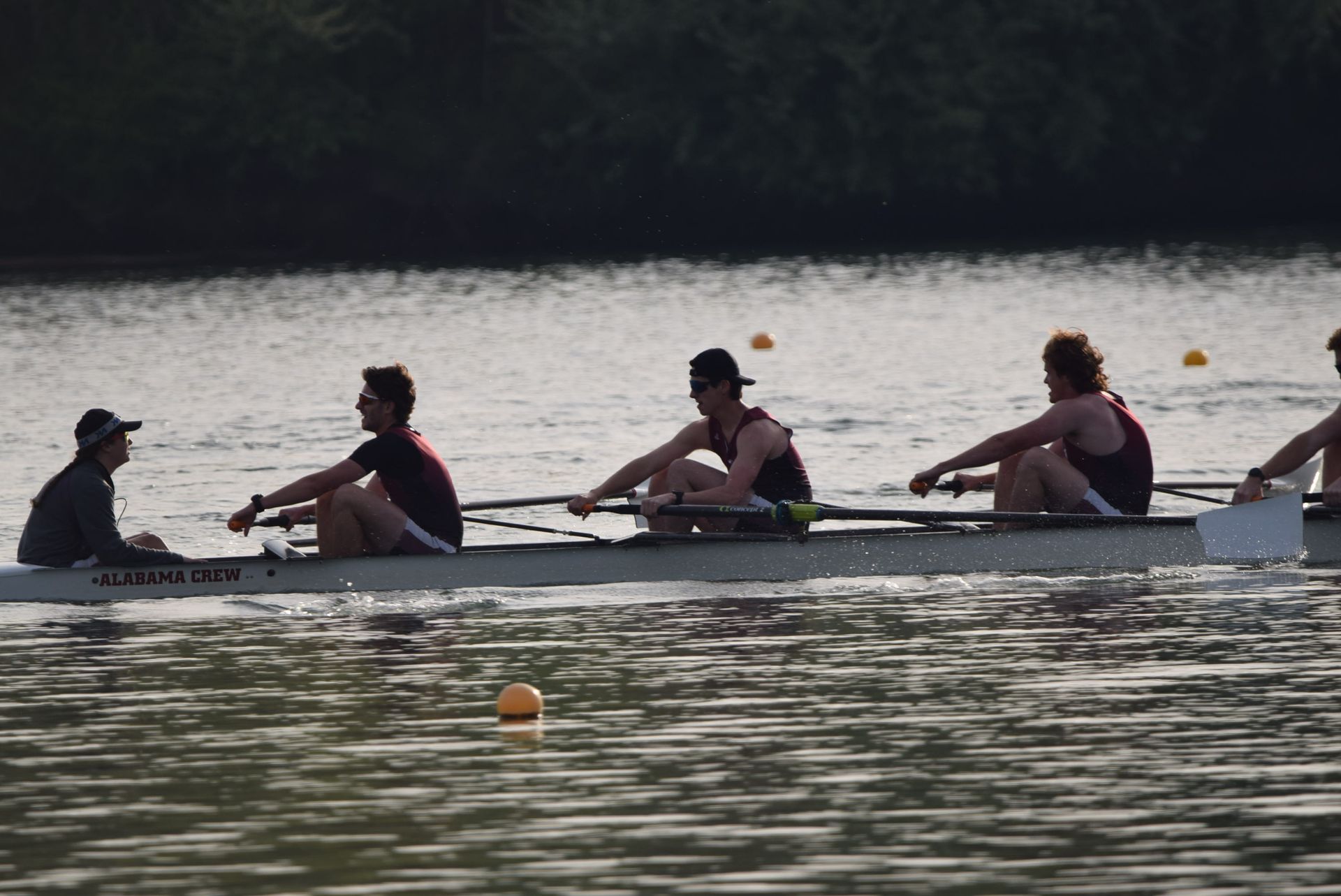 Rowing crew in a boat on a calm river.  Four rowers and a coxswain are wearing dark tops, pulling oars. 