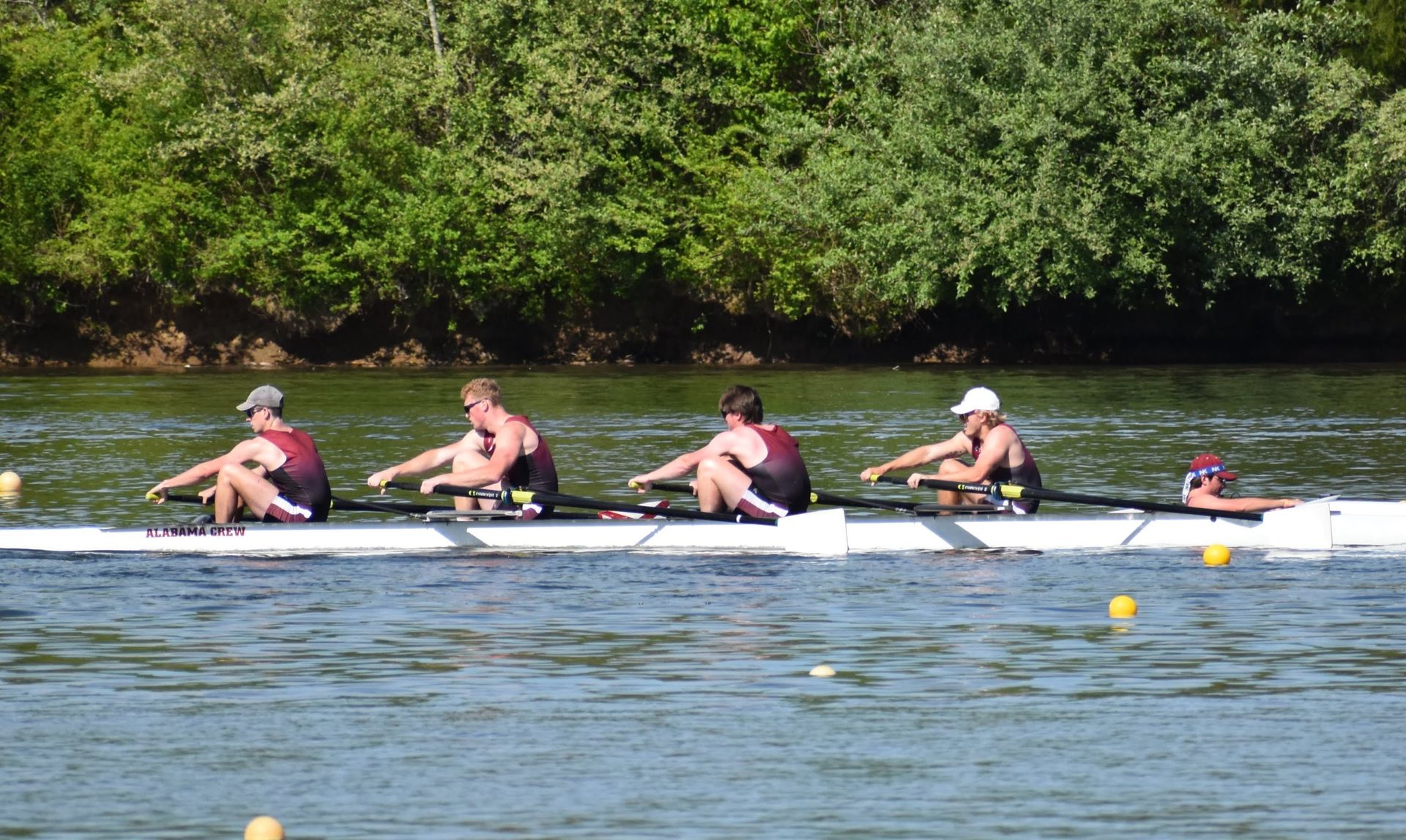 Rowing crew in a long boat on a river; athletes wearing dark uniforms, rowing in unison under a sunny sky.