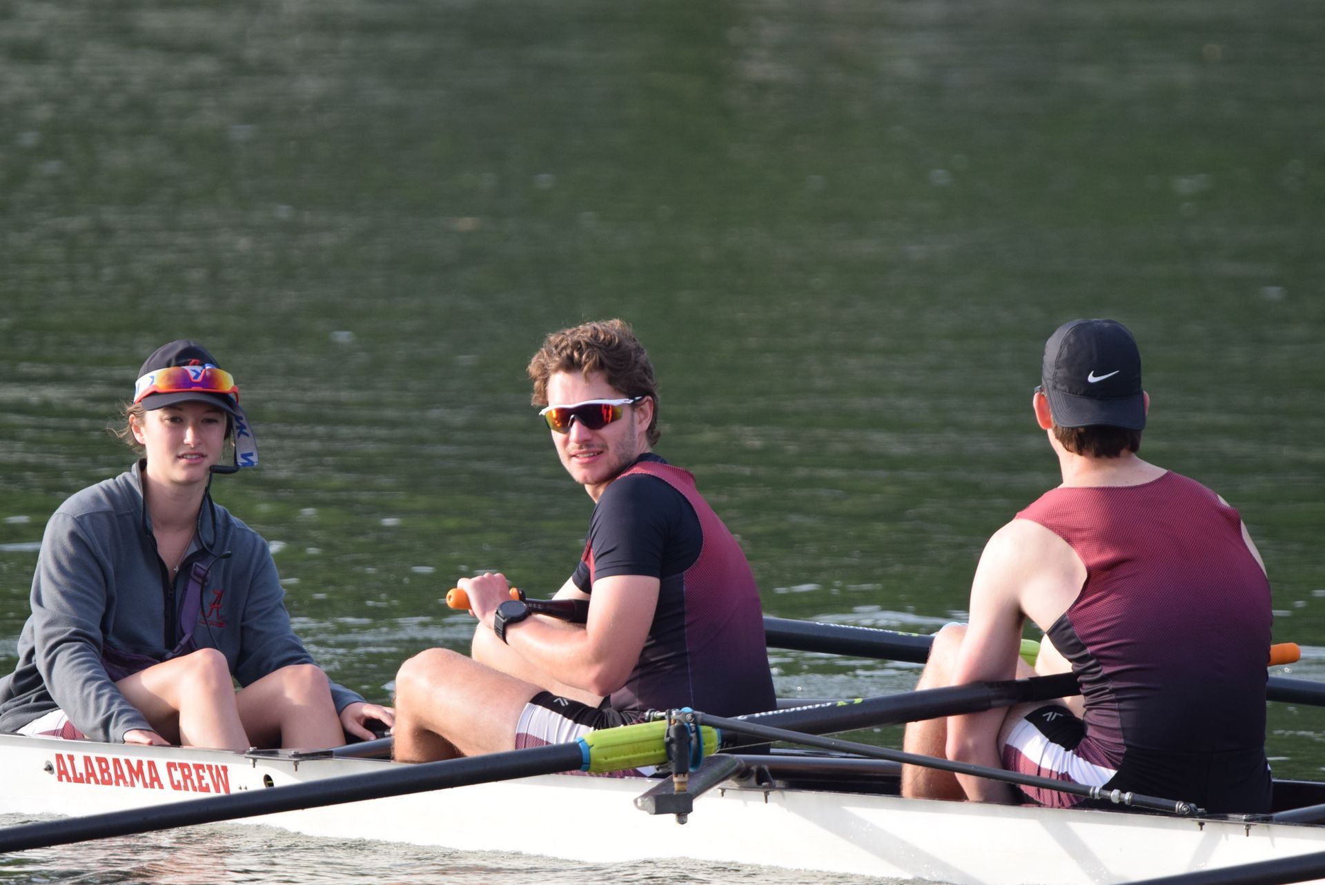 Three rowers in a boat on water; two men with sunglasses smile at the camera, another looks forward.