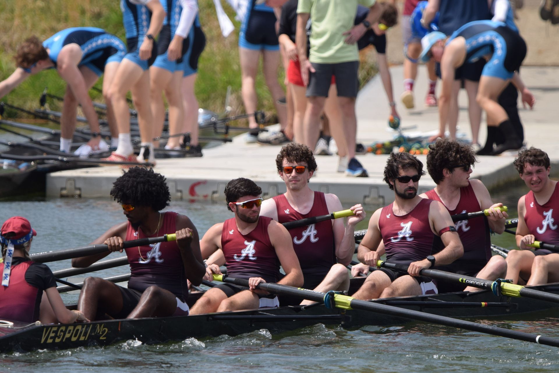 Rowing team in maroon singlets, racing on water; other teams in background.