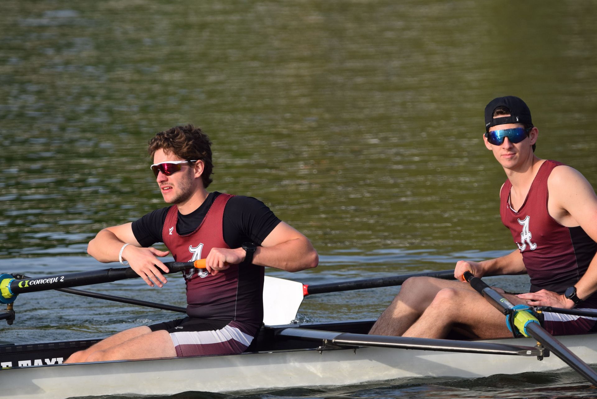 Two rowers in a boat on a lake. They wear sunglasses, maroon uniforms, and are mid-stroke. The setting is outdoors.