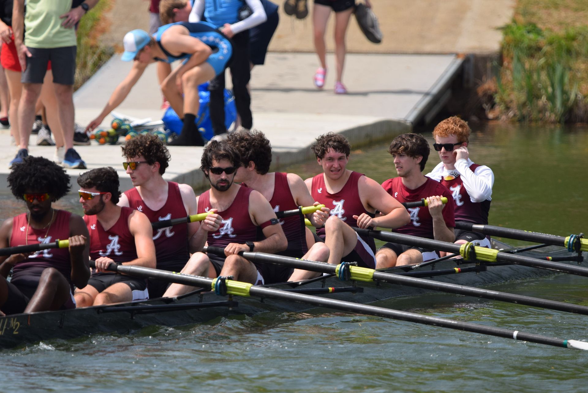 A crew team in maroon and white jerseys rows on a waterway. Spectators and support staff are in the background.