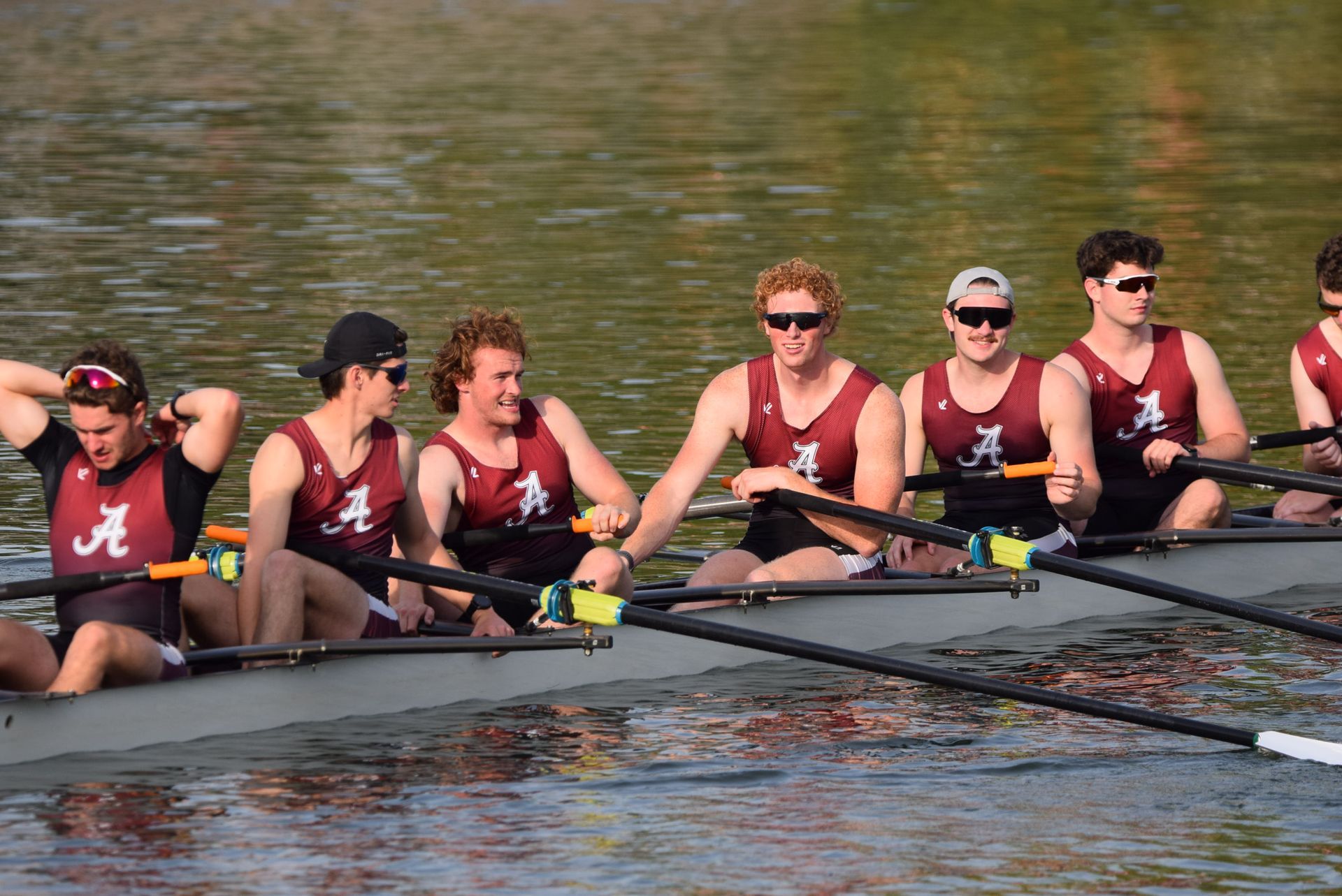 Rowing team in a boat, wearing maroon and white singlets. They are on the water.