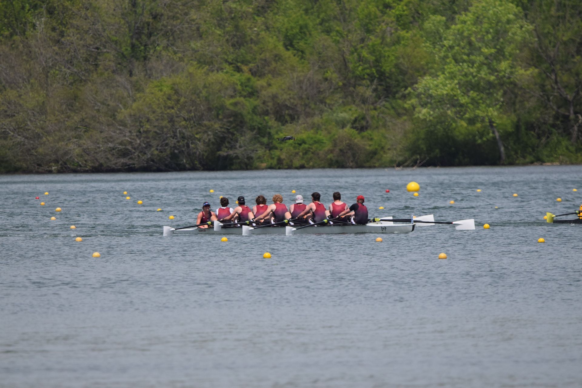 A crew team of eight rowers in maroon and white uniforms races on a lake, with trees in the background.