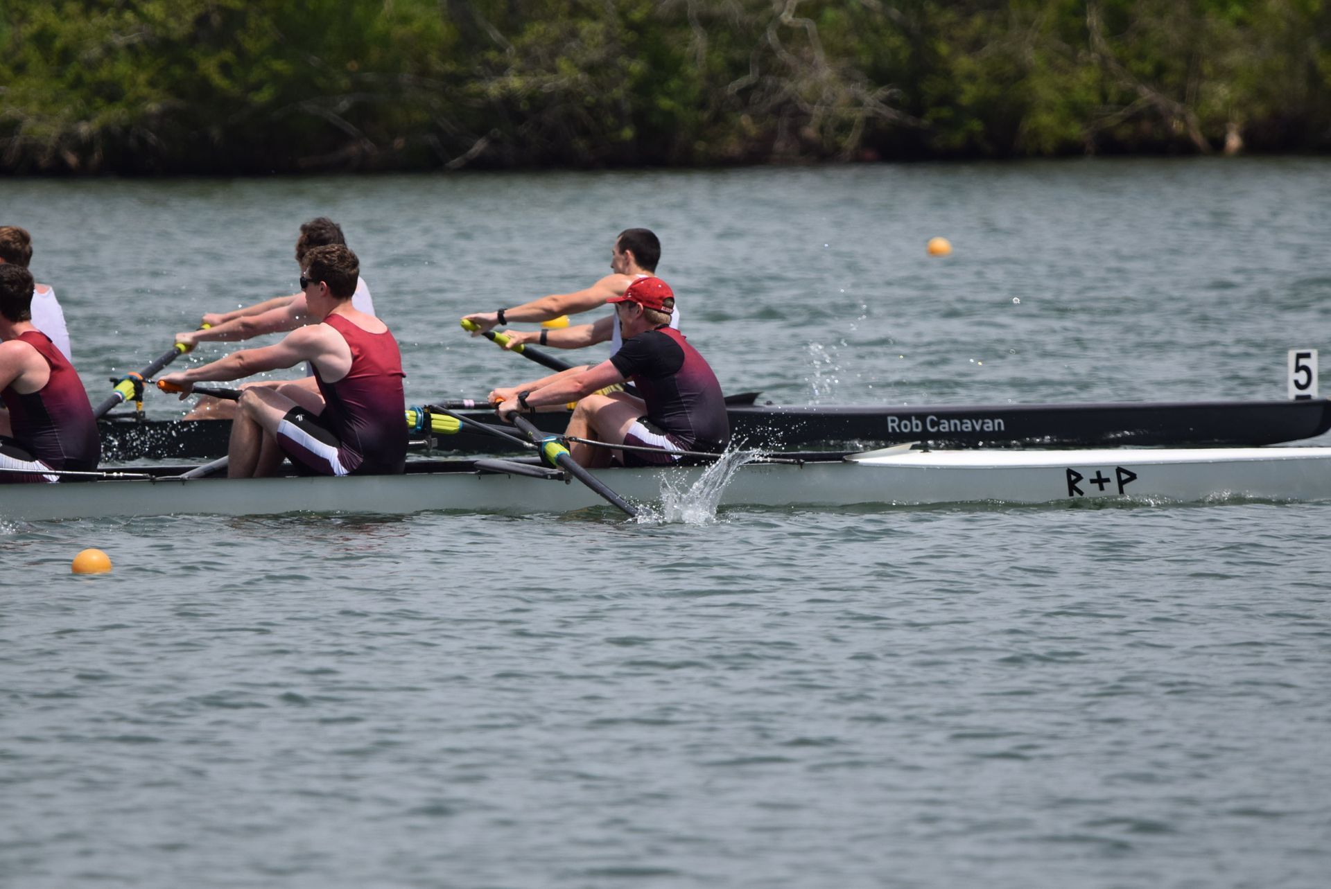 Rowers in a maroon and white boat compete on a waterway; the lead boat is splashing water.