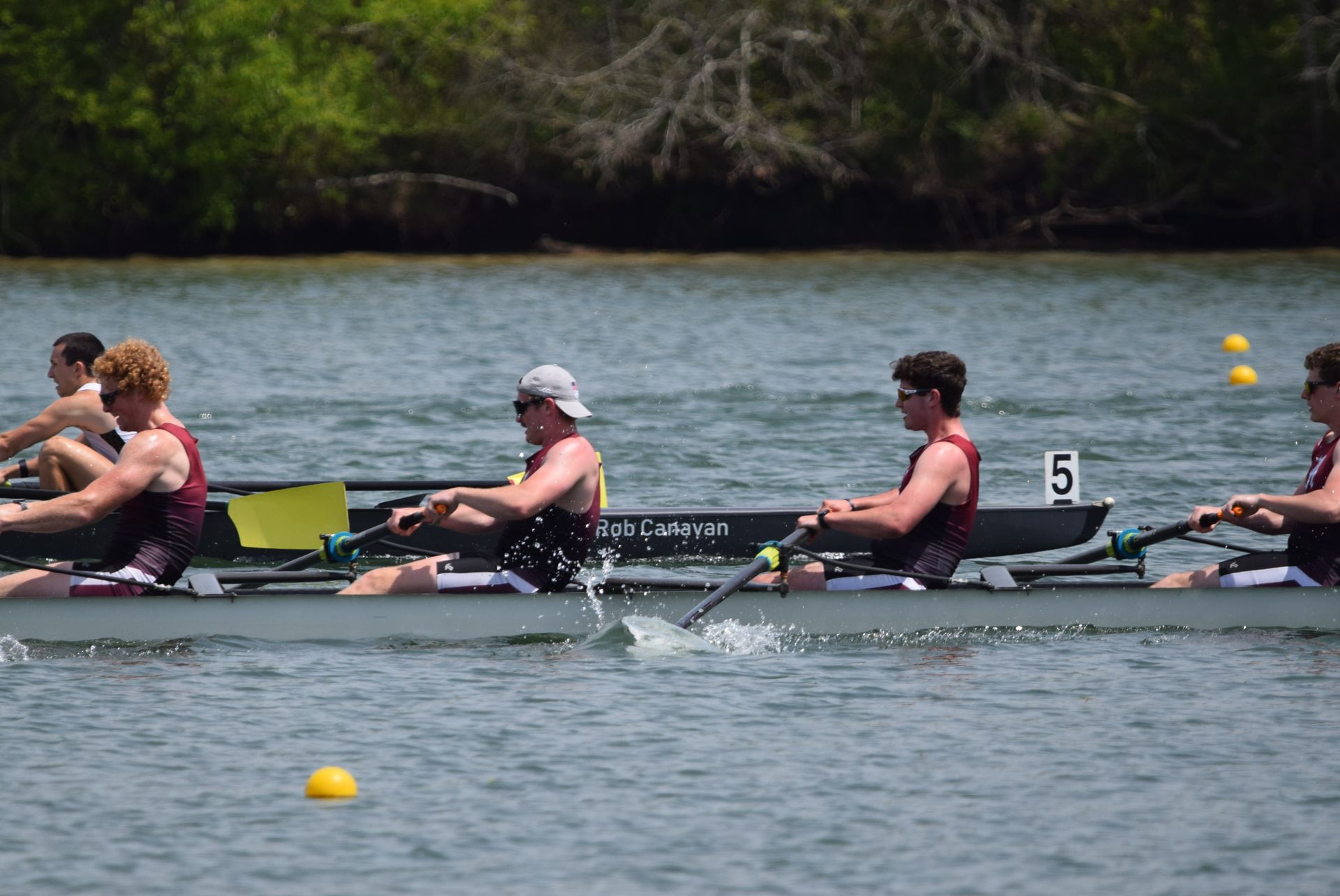 Rowing team racing in a boat on a river. Four rowers in maroon and white uniforms are visible; the boat is black.