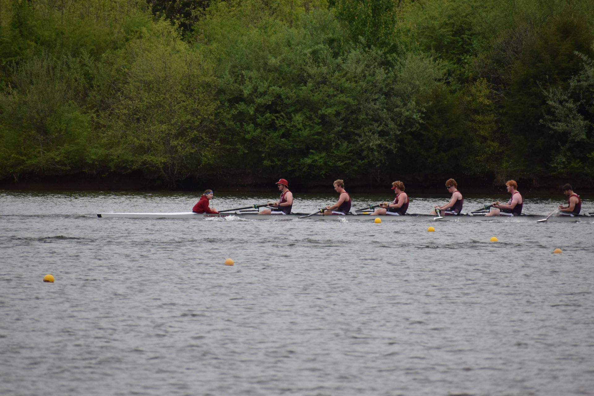 Rowing team on a lake, six athletes in dark uniforms and one coxswain in red. They row towards the viewer.