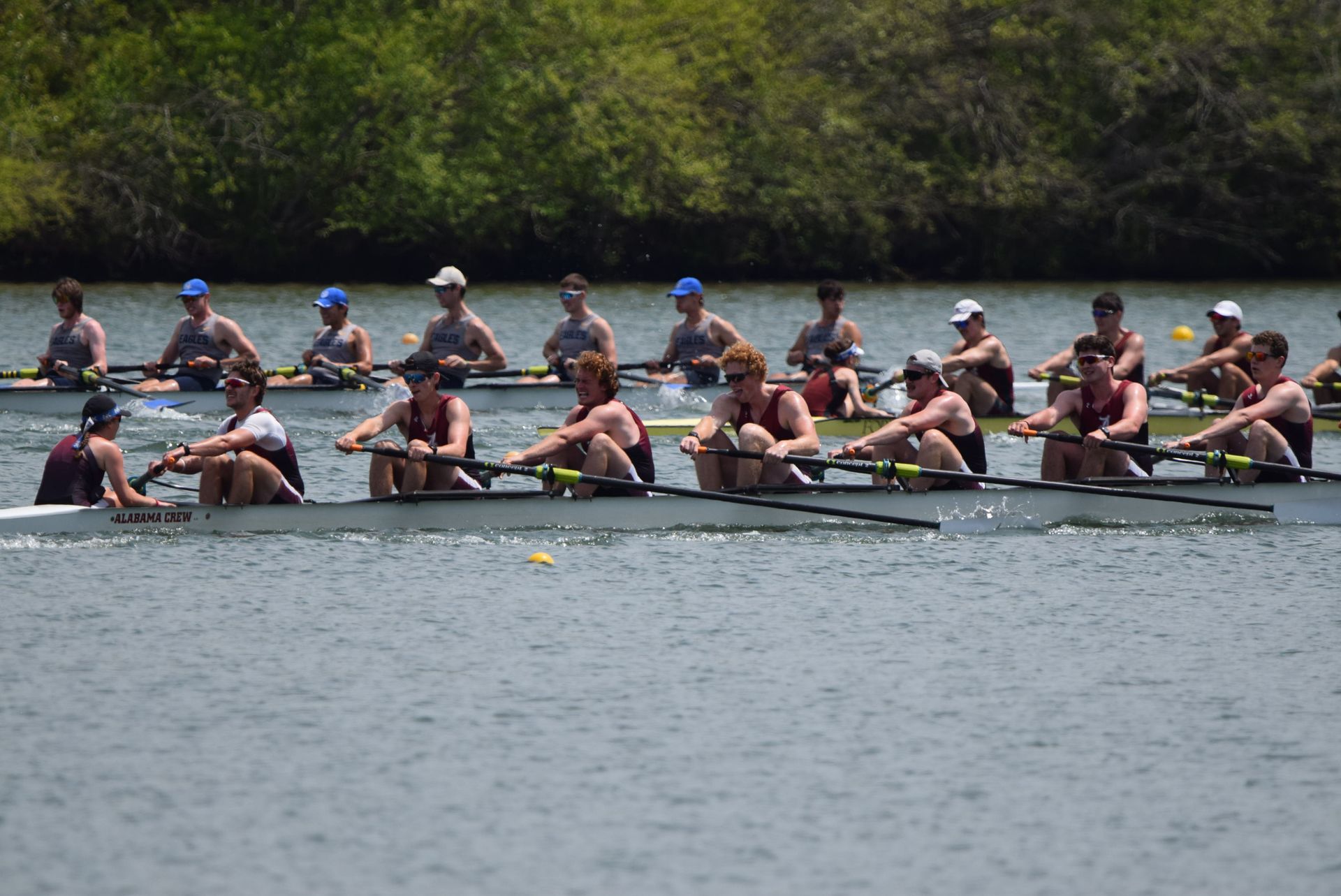 Rowing teams in long boats competing on a river, under a blue sky. Athletes in team uniforms are rowing.