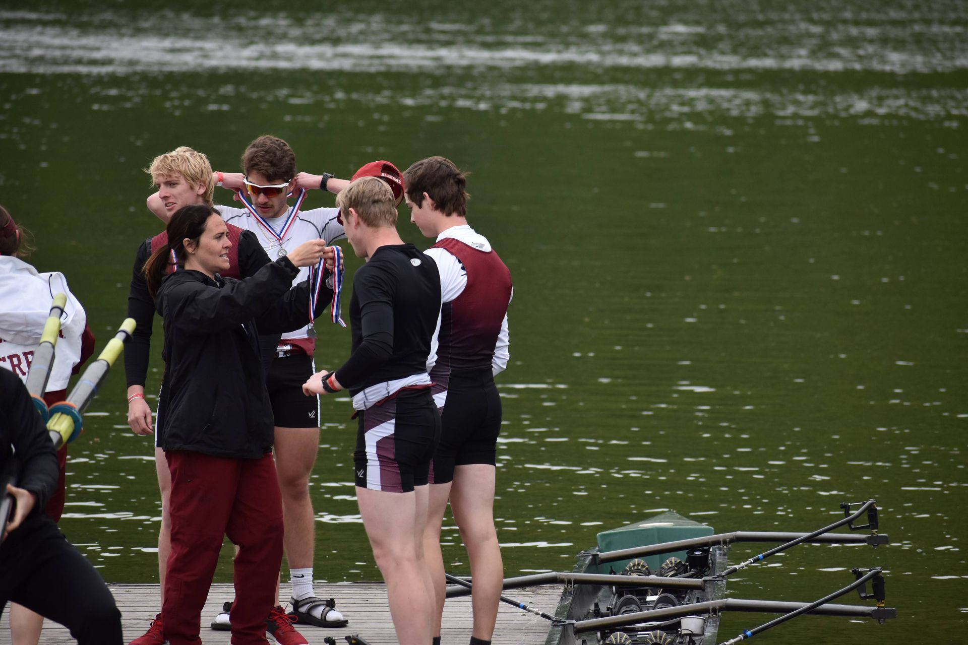 A woman places a medal on a rower's neck on a wooden dock. Several male rowers stand nearby, in a lakeside setting.