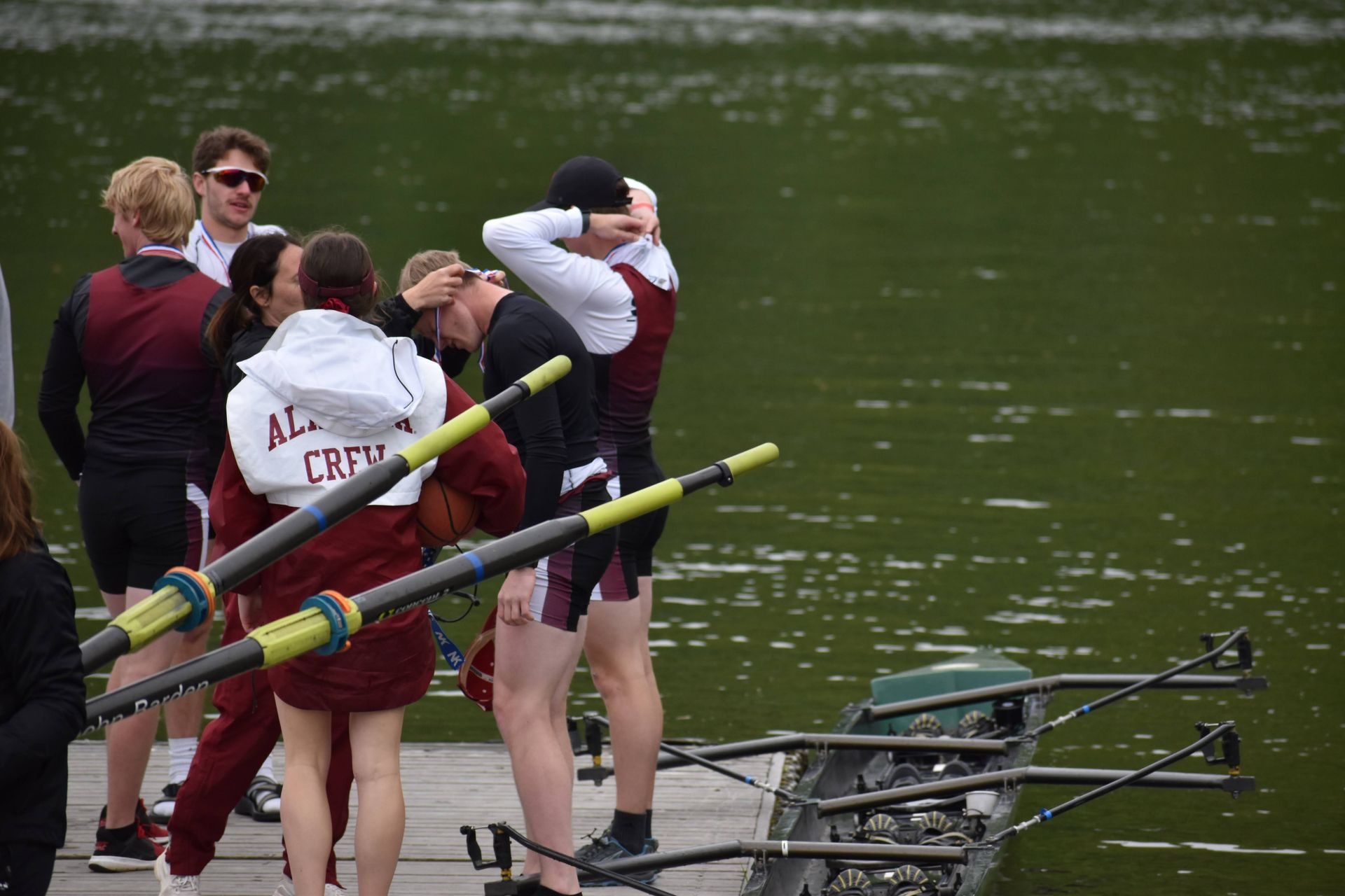 Rowing team on a dock. Some athletes embrace and stretch, while others hold oars near a boat.