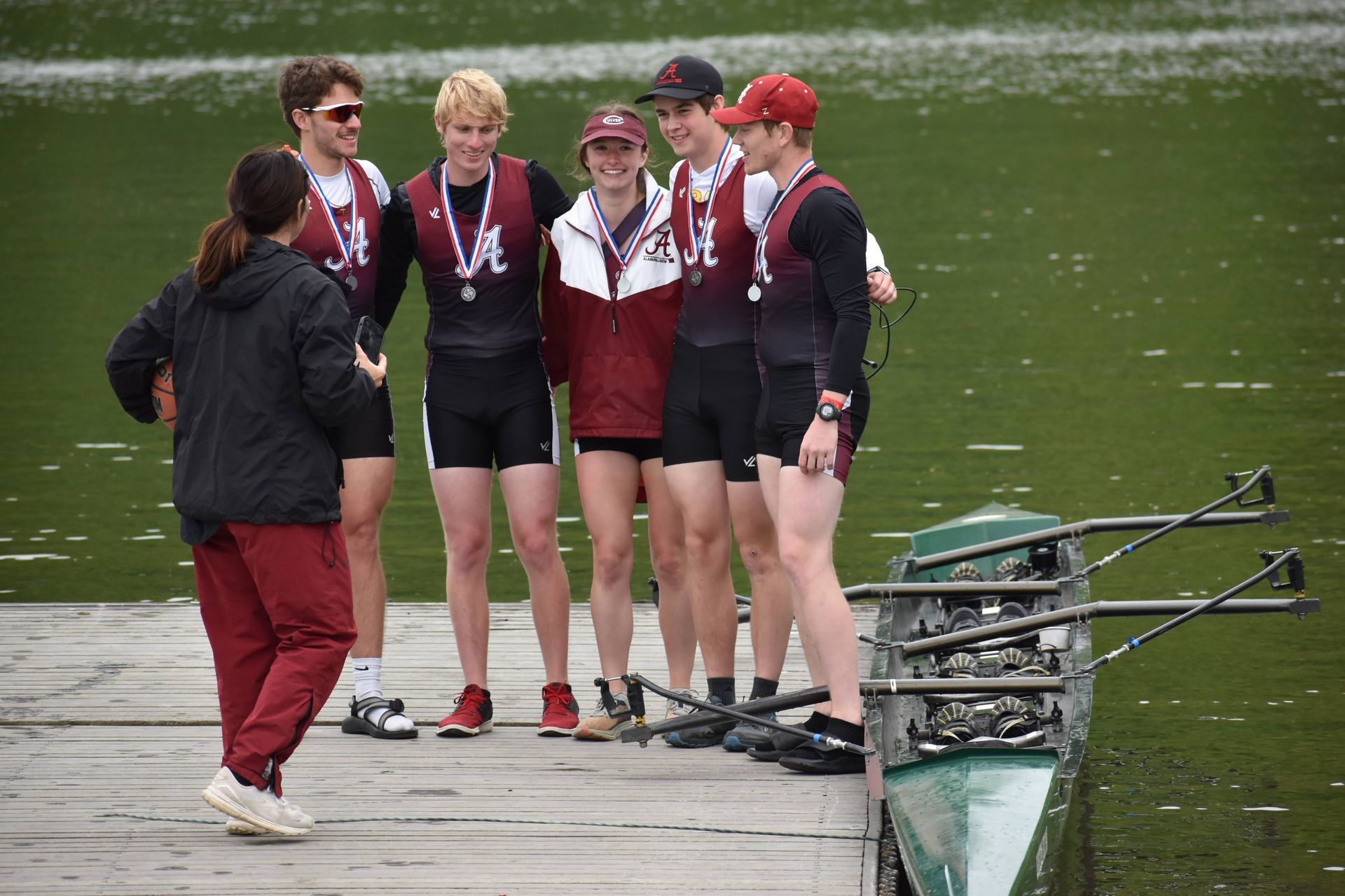 Rowing team with medals stands on a dock next to their boat, receiving instruction from a coach. They wear maroon and black.
