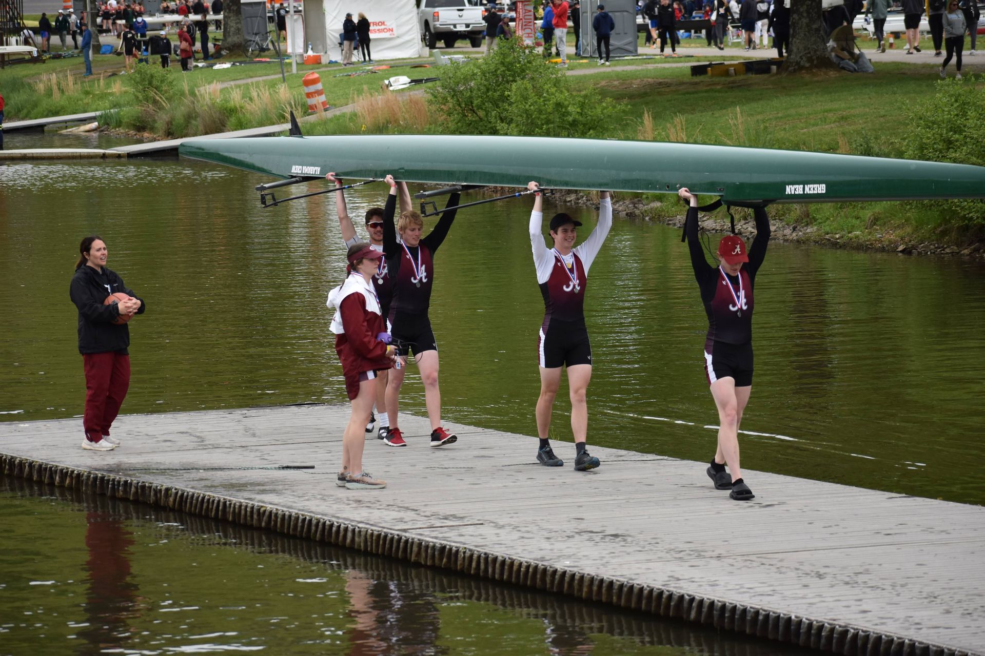 Rowing team carrying a long, green boat over their heads on a dock. Spectators and water in background.