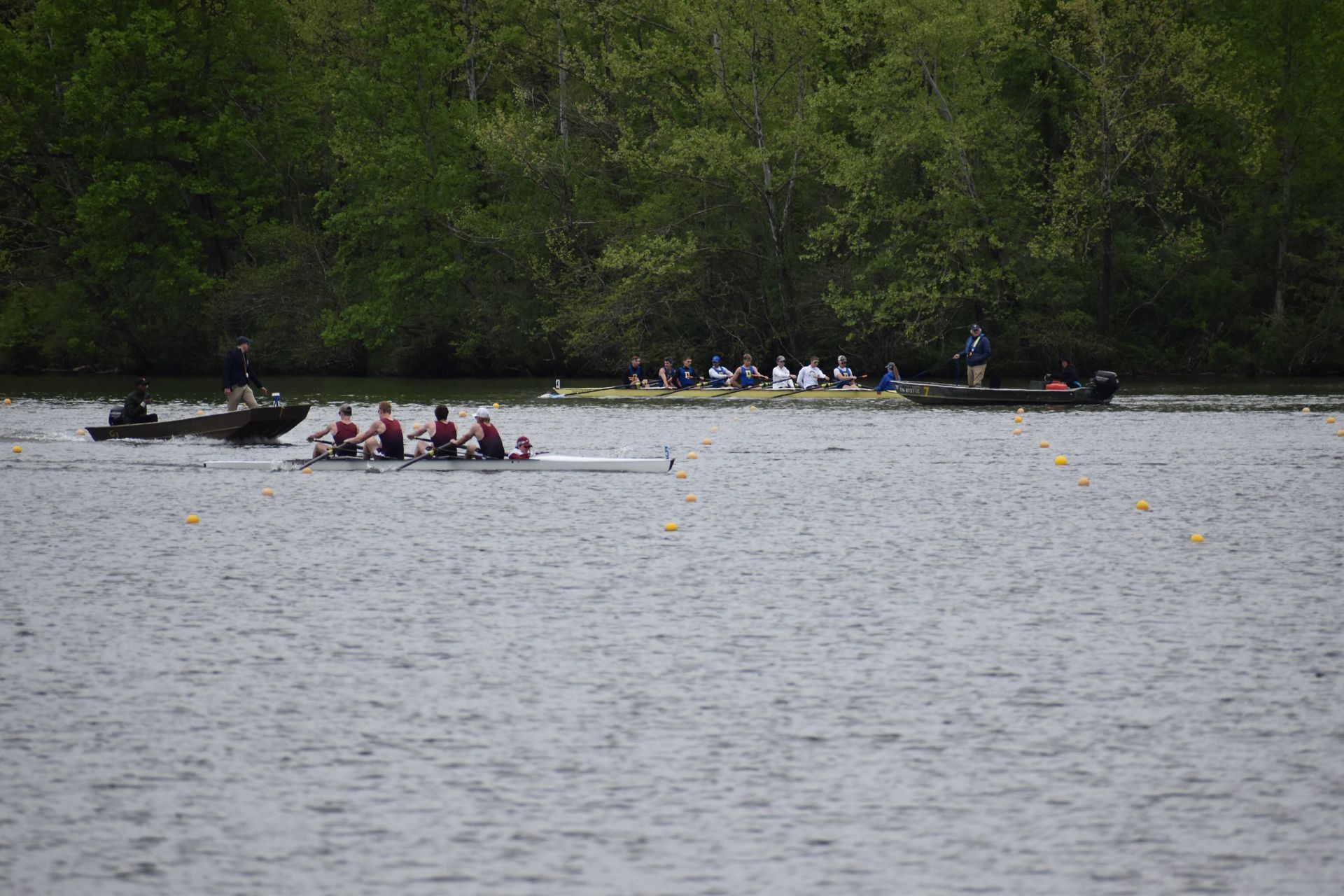 Rowing teams compete on a lake, with several boats racing. Trees line the background, and a boat with a motor observes.