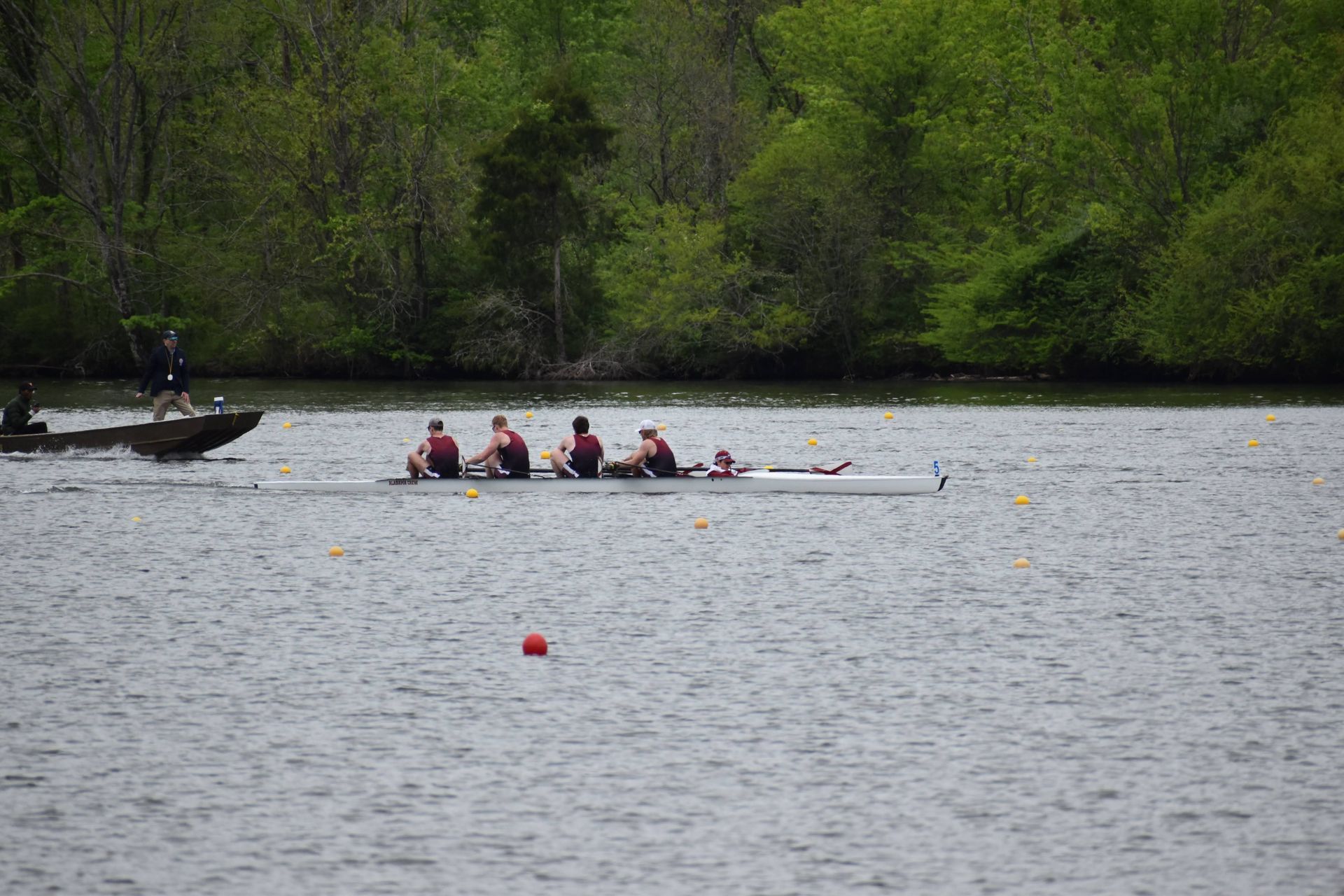 Rowing team in a long boat on a river with a motorboat nearby. The rowers are pulling the oars in unison.