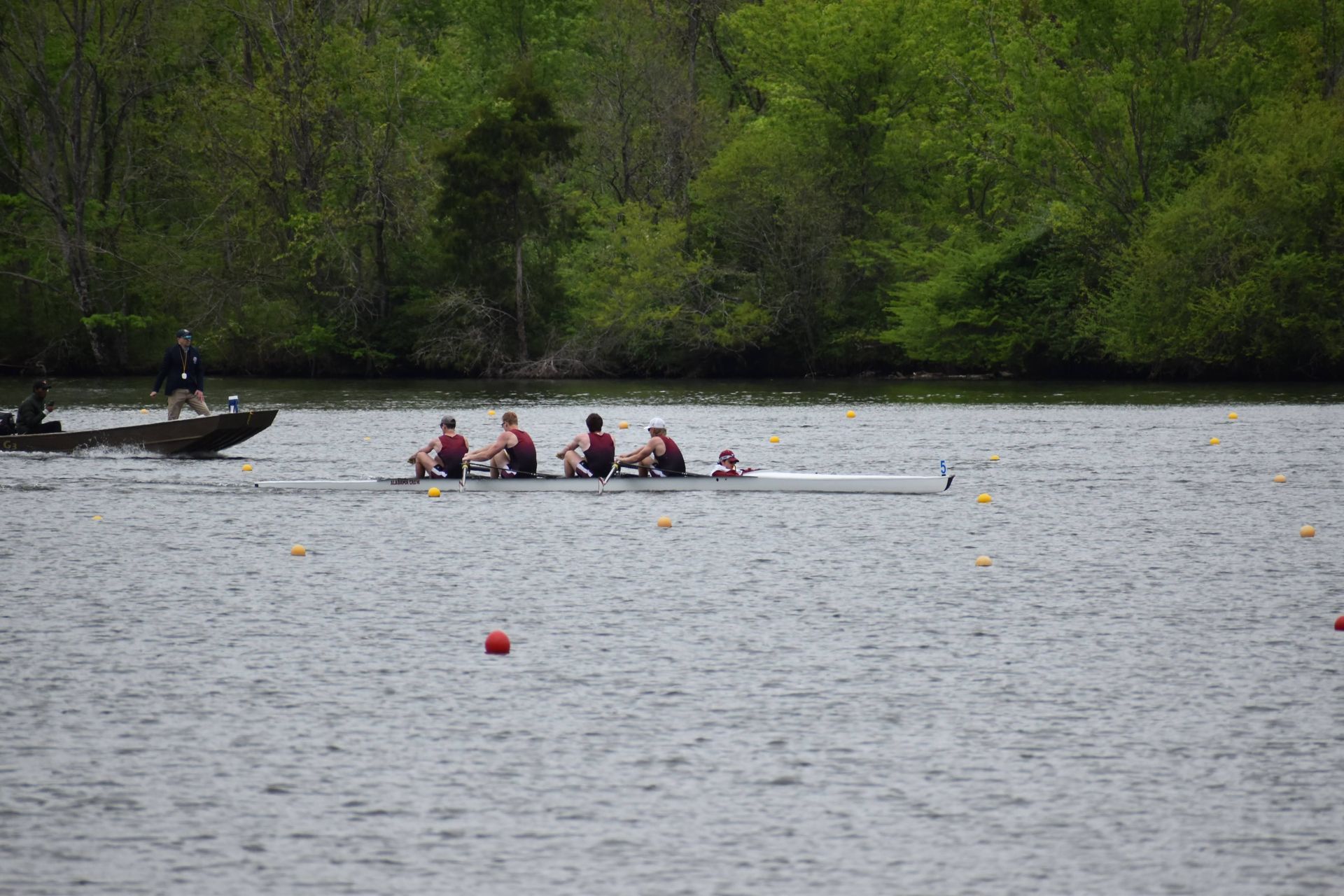 Rowing crew on a lake, four athletes in a boat, a motorboat nearby, trees in the background.