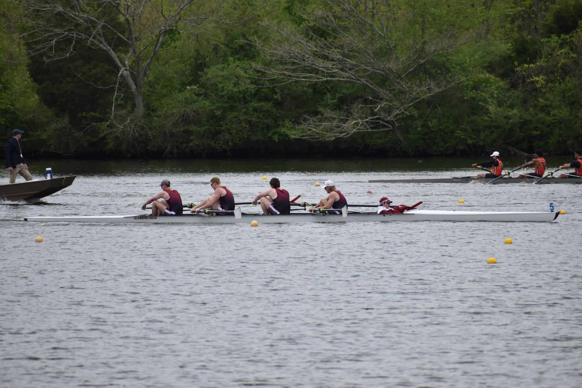 Rowing team in a four-person shell on a river, with another boat in the distance. Dark green and brown trees line the water.