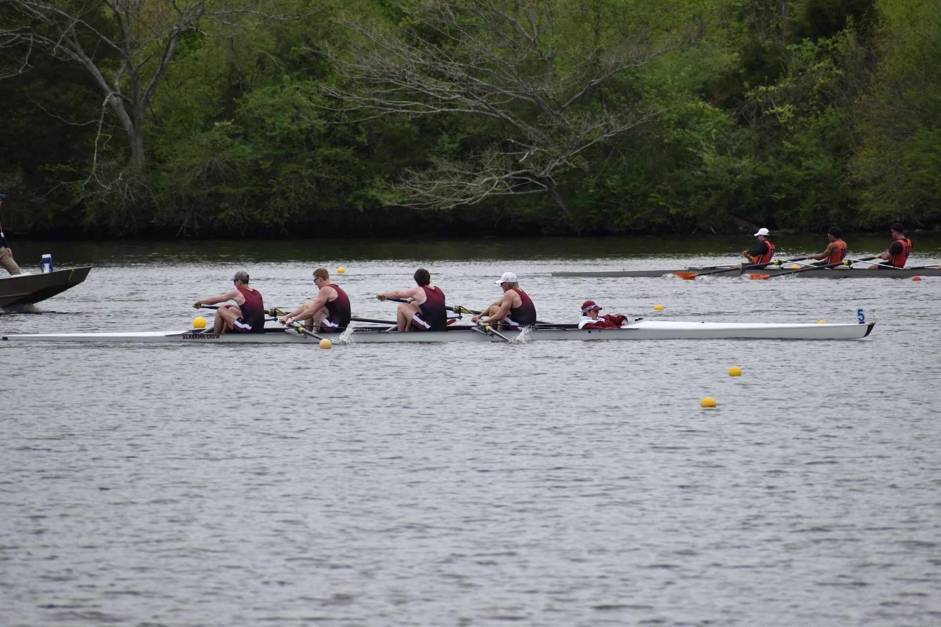 Rowing crew on a river, dark uniforms. Crew boat in foreground, other boats and shoreline in the distance.