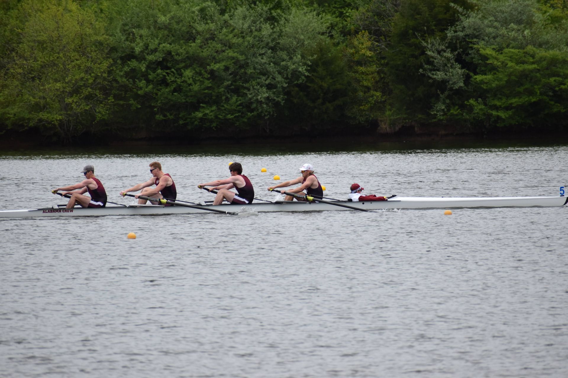 Four rowers in a boat on a river, dark blue uniforms, rowing towards the right; trees line the far bank.