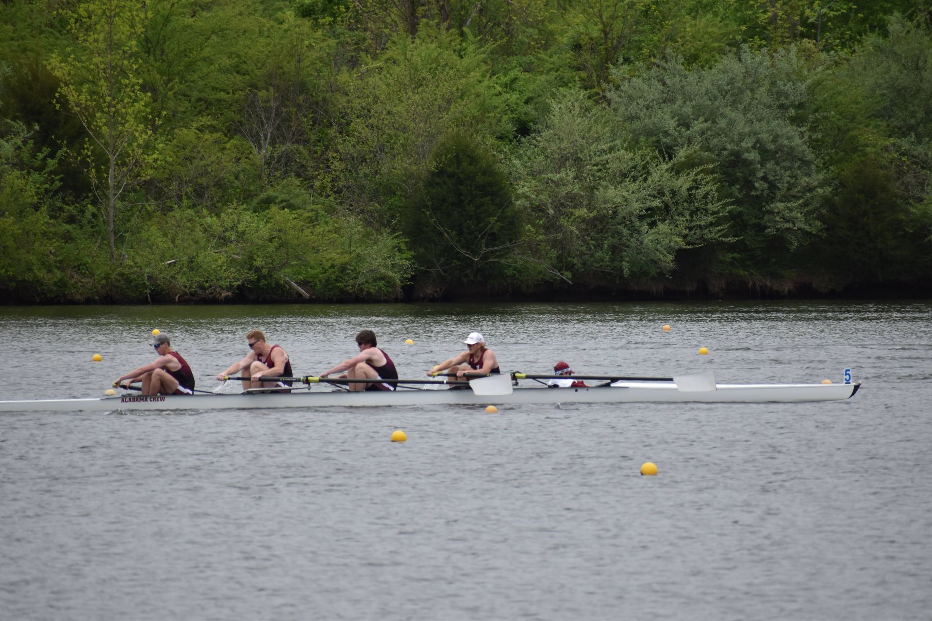 Rowing crew on a lake, four athletes in dark uniforms pulling oars. Trees line the riverbank.